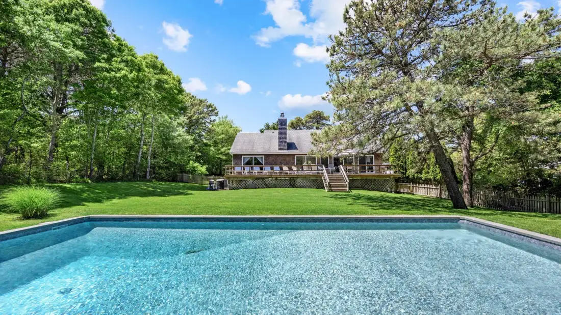 An outdoor swimming pool faces a house across a grassy lawn.