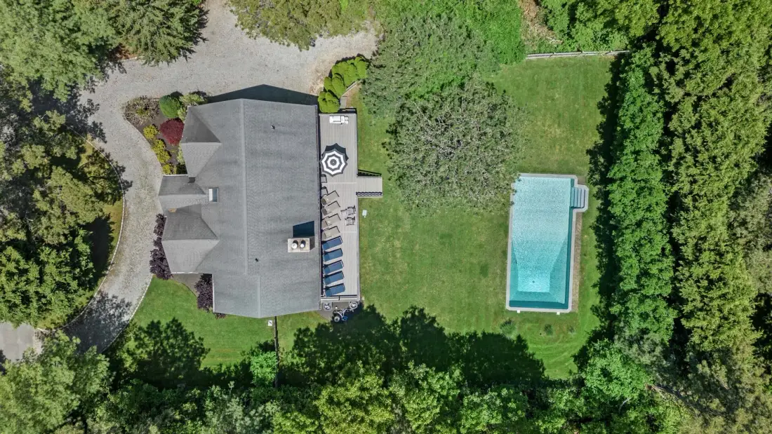An aerial view shows a house, a lawn, and a rectangular swimming pool surrounded by trees.