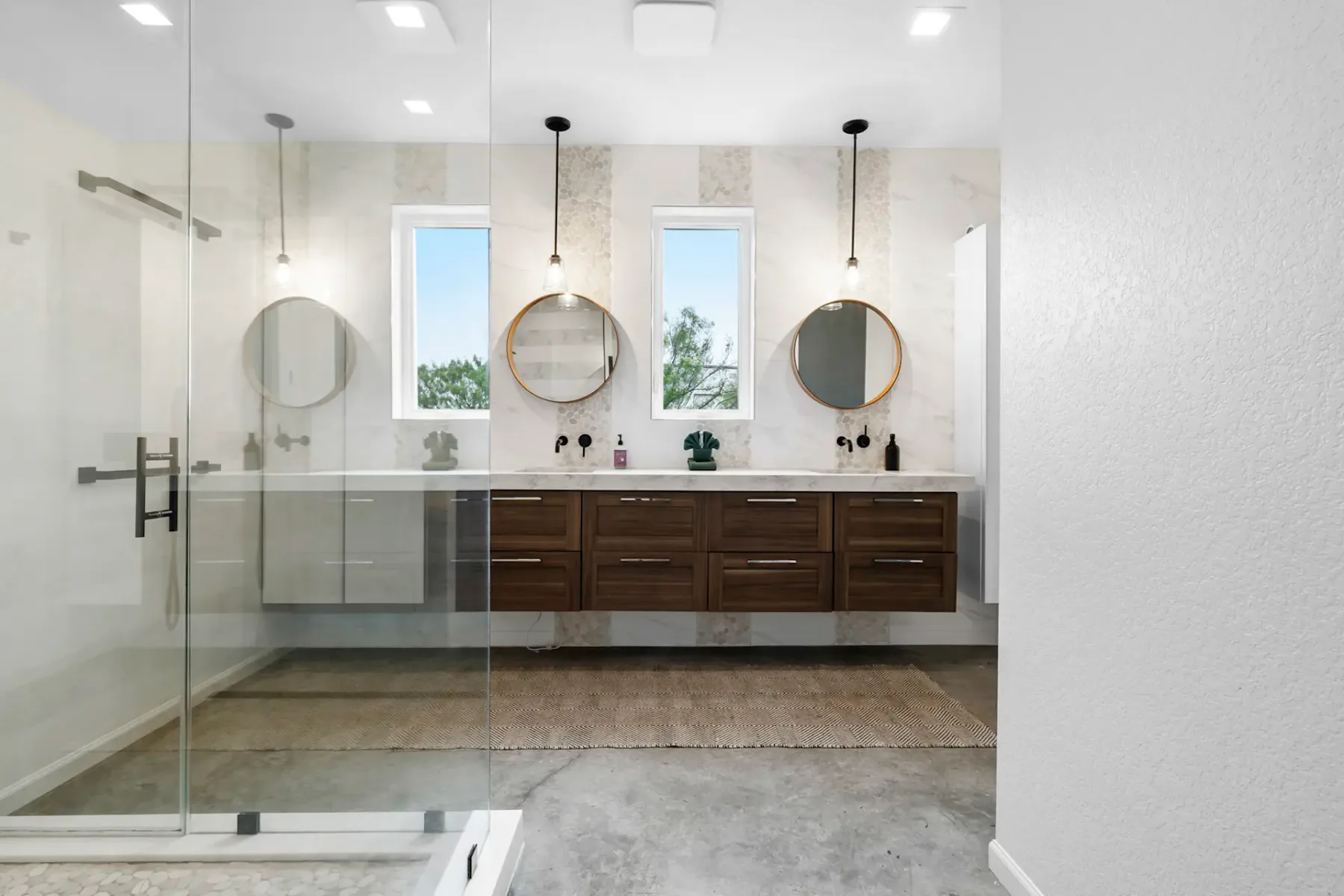 Modern bathroom with a glass-enclosed shower, double vanity with wooden cabinets, circular mirrors, and a concrete floor.