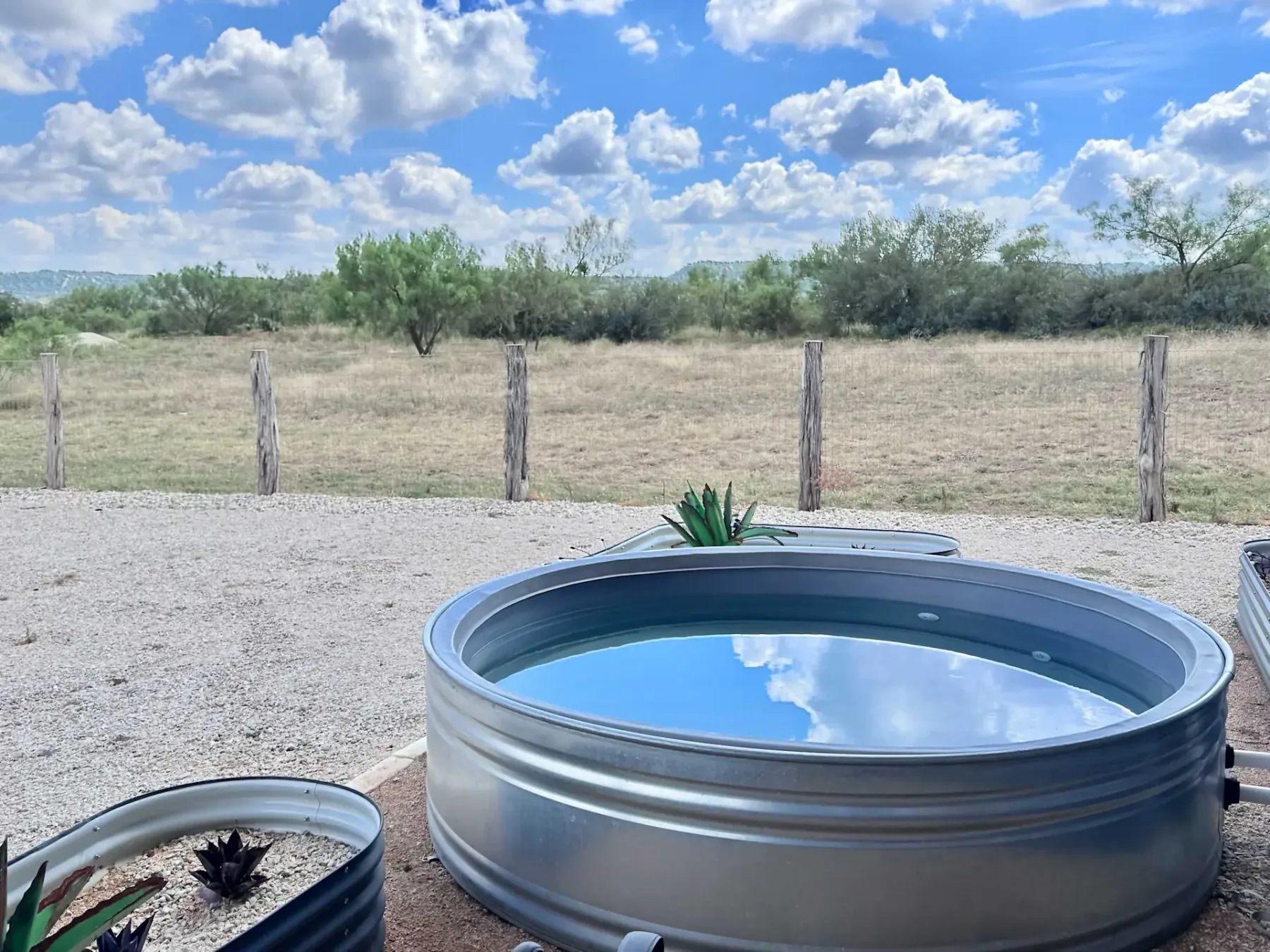 Metal tub with water reflecting a cloudy blue sky, set in a gravel area next to a dry field, wooden fence, and scattered plants.