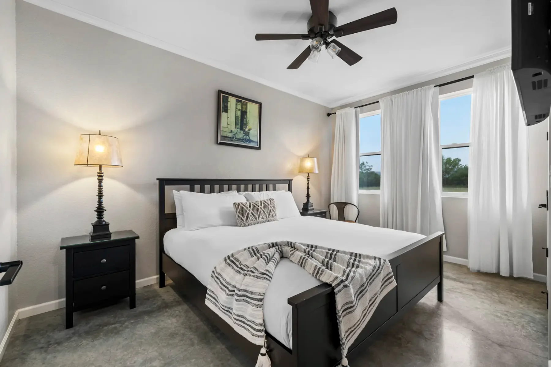 A bright bedroom with a neatly made bed, patterned pillows, and a striped blanket. White curtains cover the windows, and a ceiling fan is above.