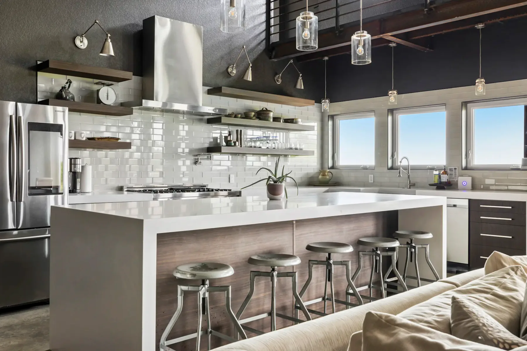 Modern kitchen with a white island, metal stools, stainless steel appliances, pendant lights, and large windows for natural light.