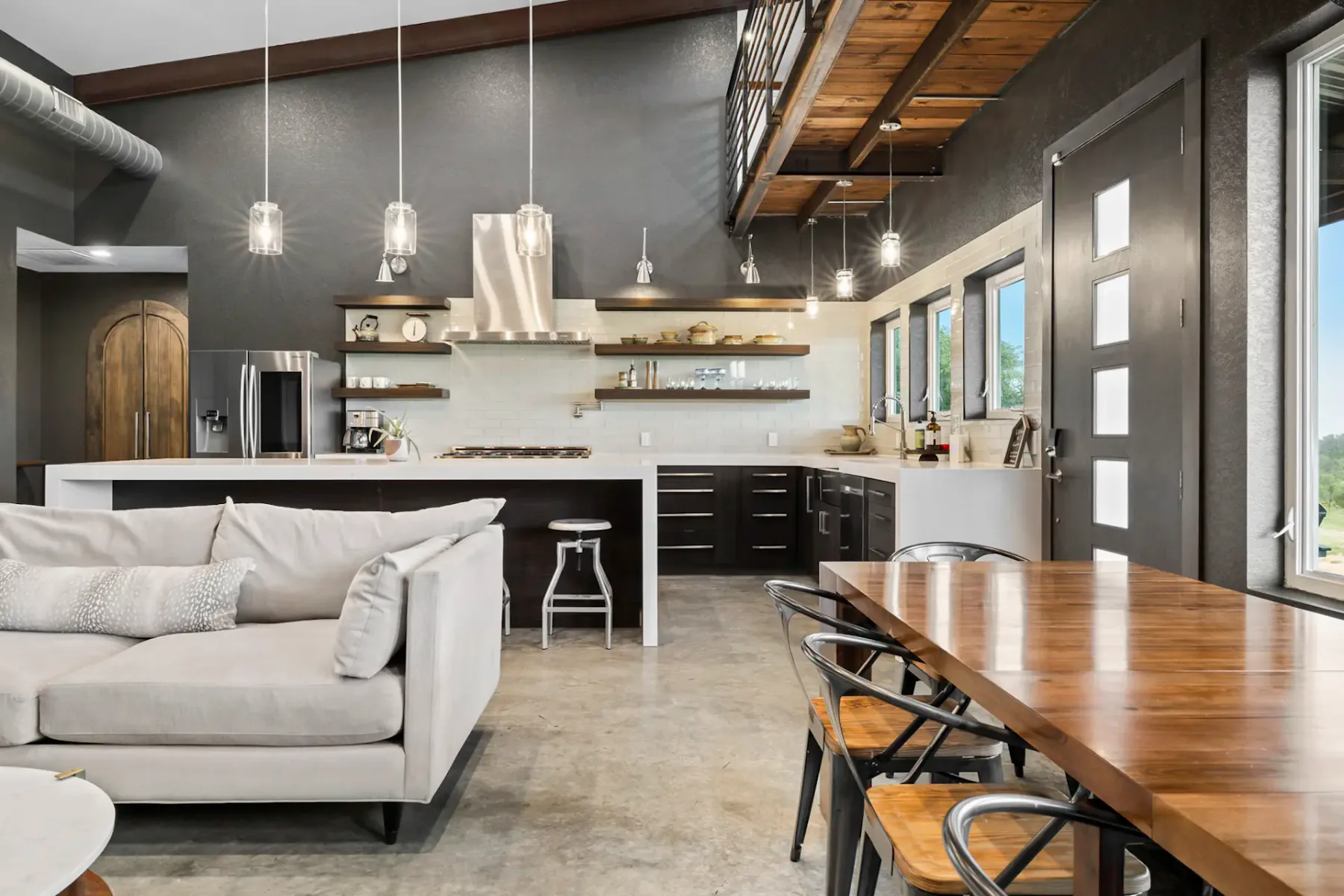 Modern open-concept kitchen and living area with high ceilings, pendant lights, a beige sofa, wooden dining table, and stainless steel appliances.