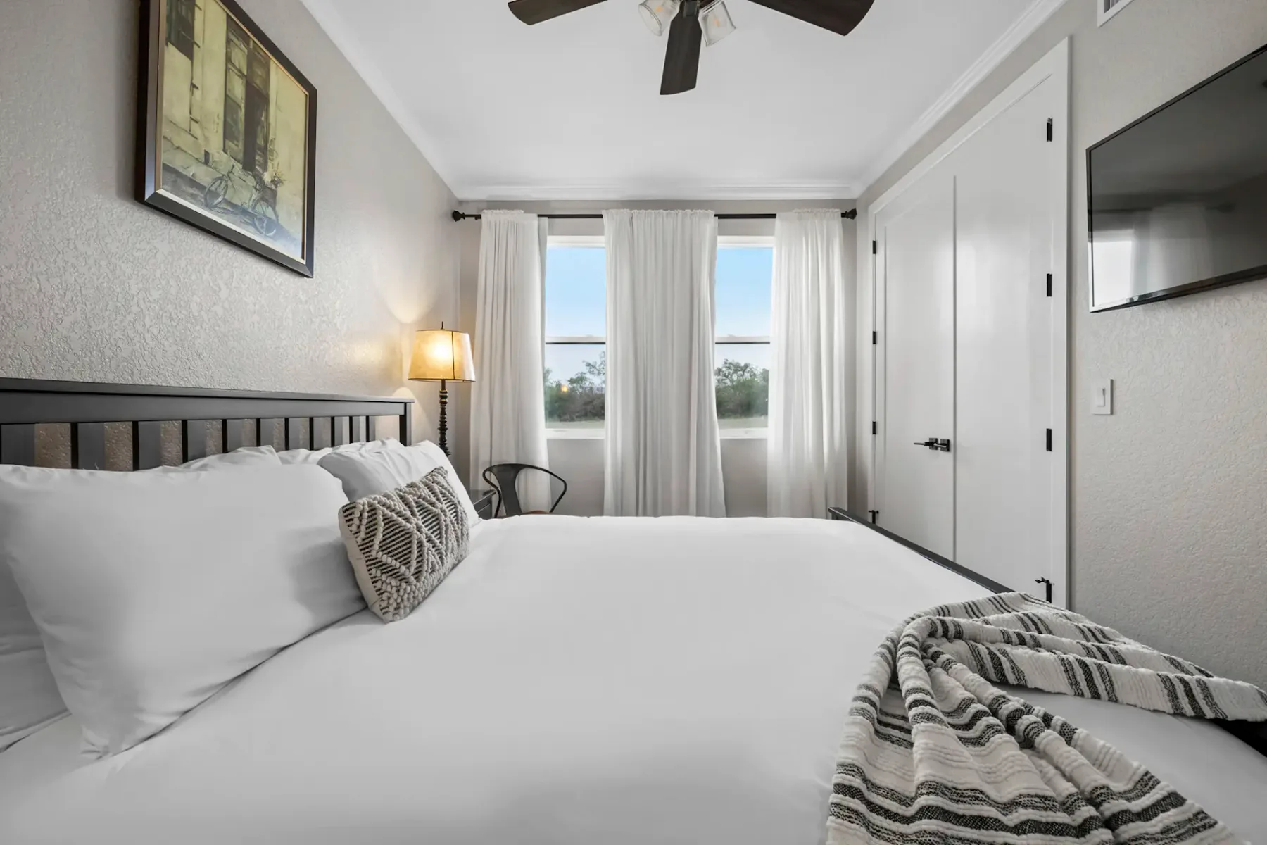 A bright bedroom with a neatly made bed, patterned pillows, and a striped blanket. White curtains cover the windows, and a ceiling fan is above.