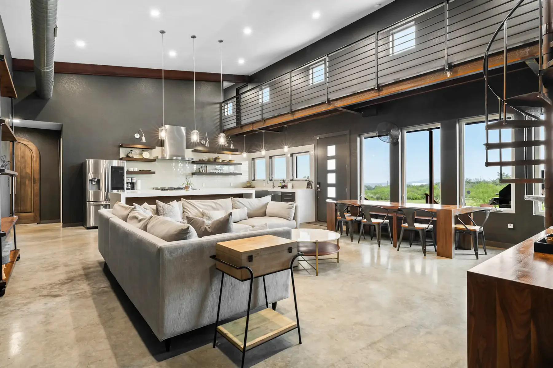 Spacious modern loft with a gray sectional sofa, spiral staircase, high ceiling, large windows, and a mix of industrial and rustic decor.
