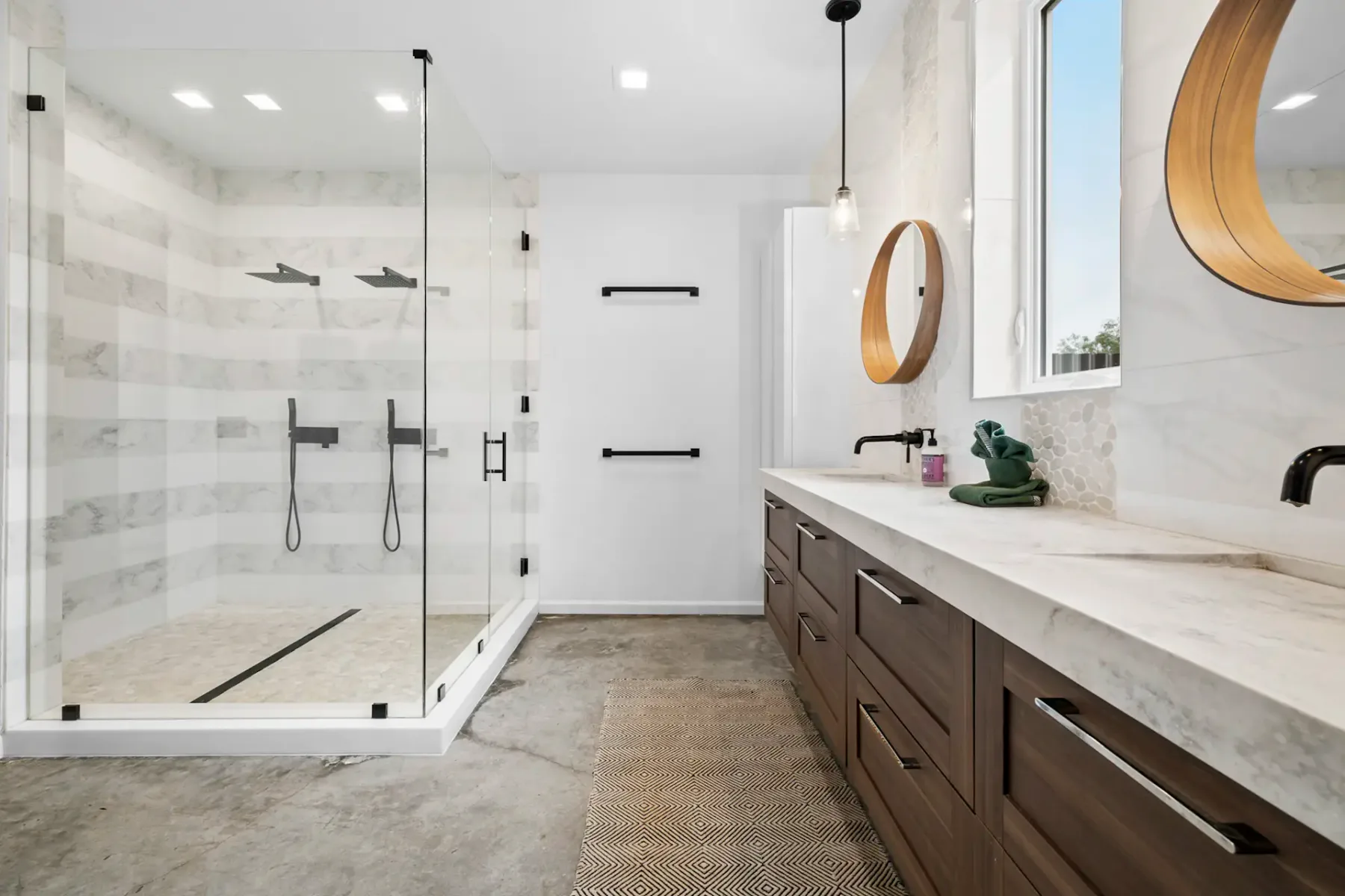 Modern bathroom with a glass-enclosed shower, double vanity with wooden cabinets, circular mirrors, and a concrete floor.