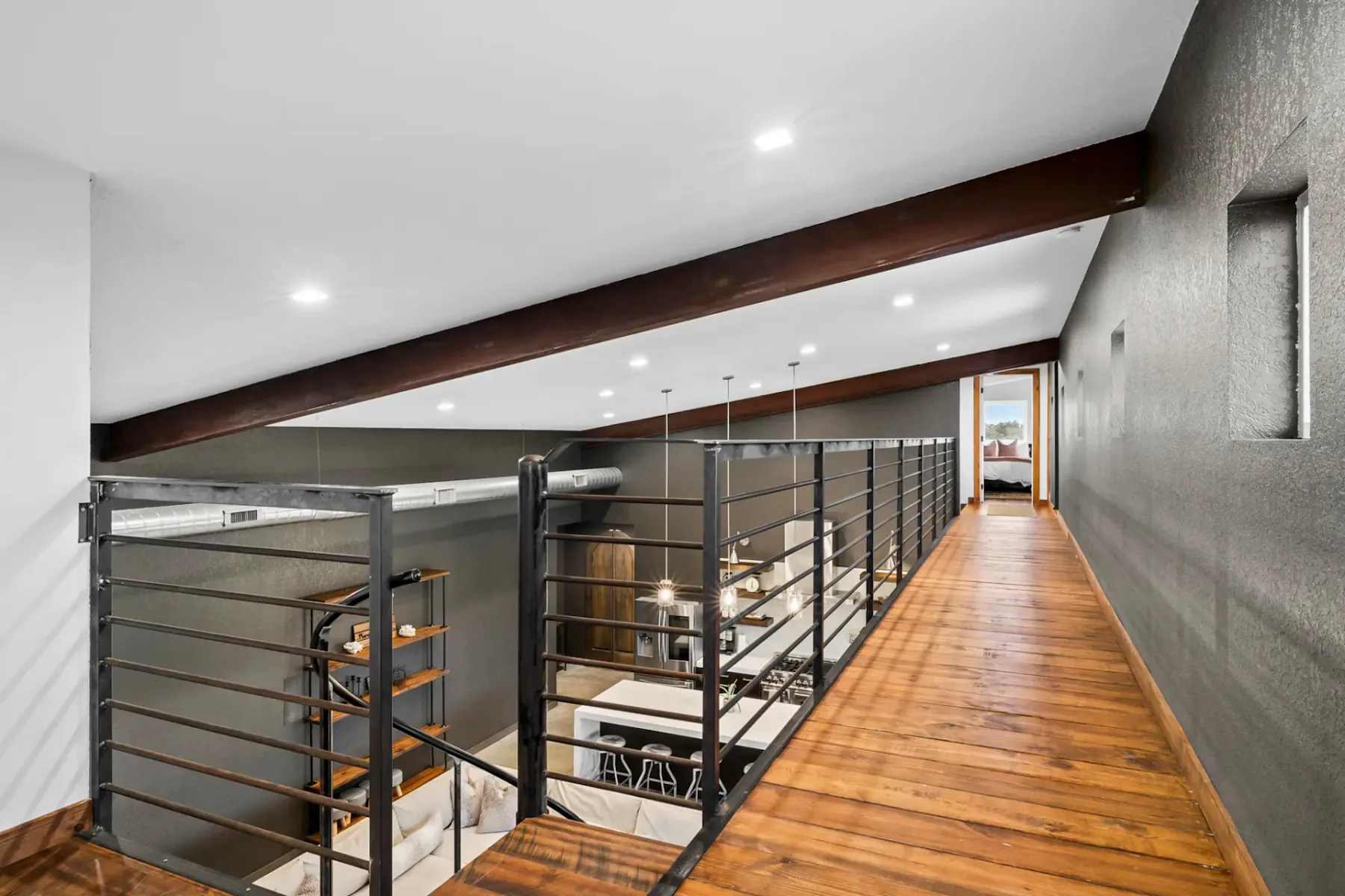Modern loft with wooden flooring, metal railings, and exposed beams, overlooking a contemporary living area with pendant lights and white walls.