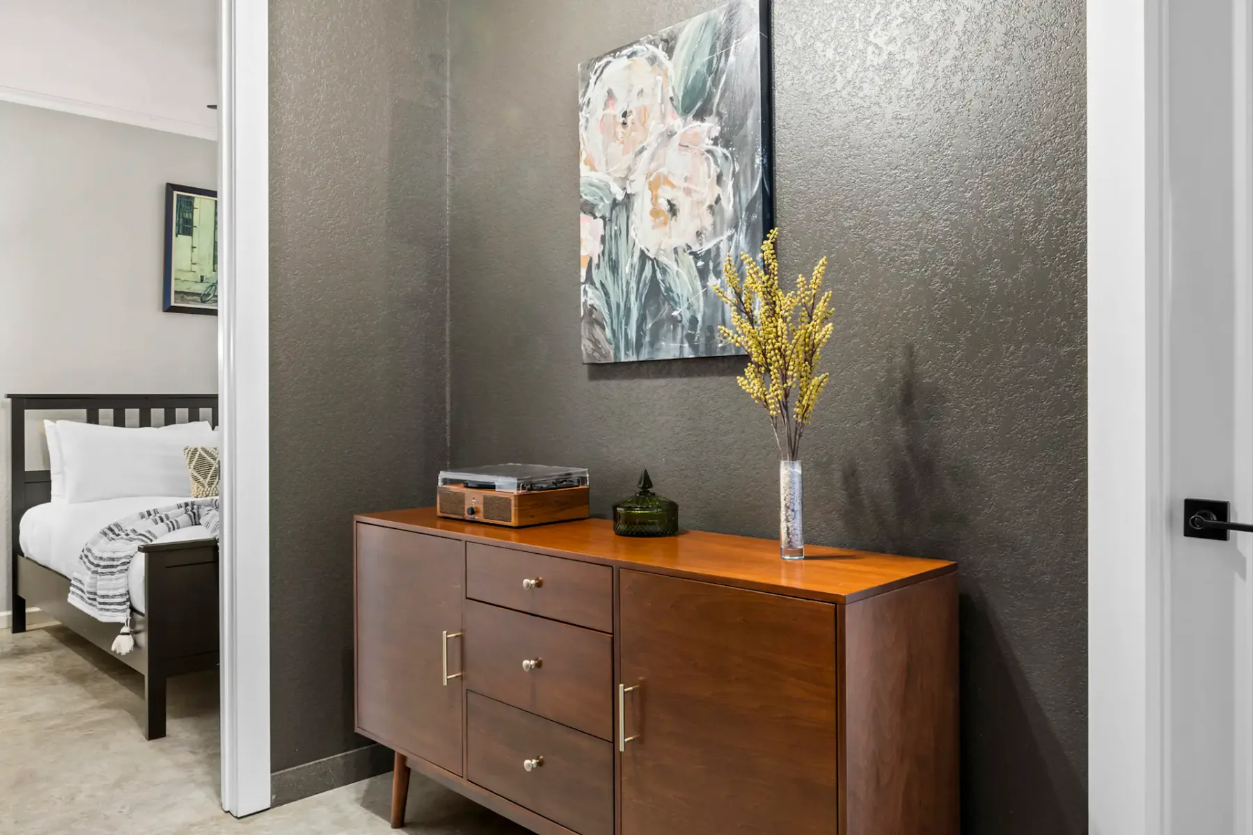 Wooden sideboard with a vase of yellow flowers, a turntable, and a painting above. Bedroom visible through an open door on the left.