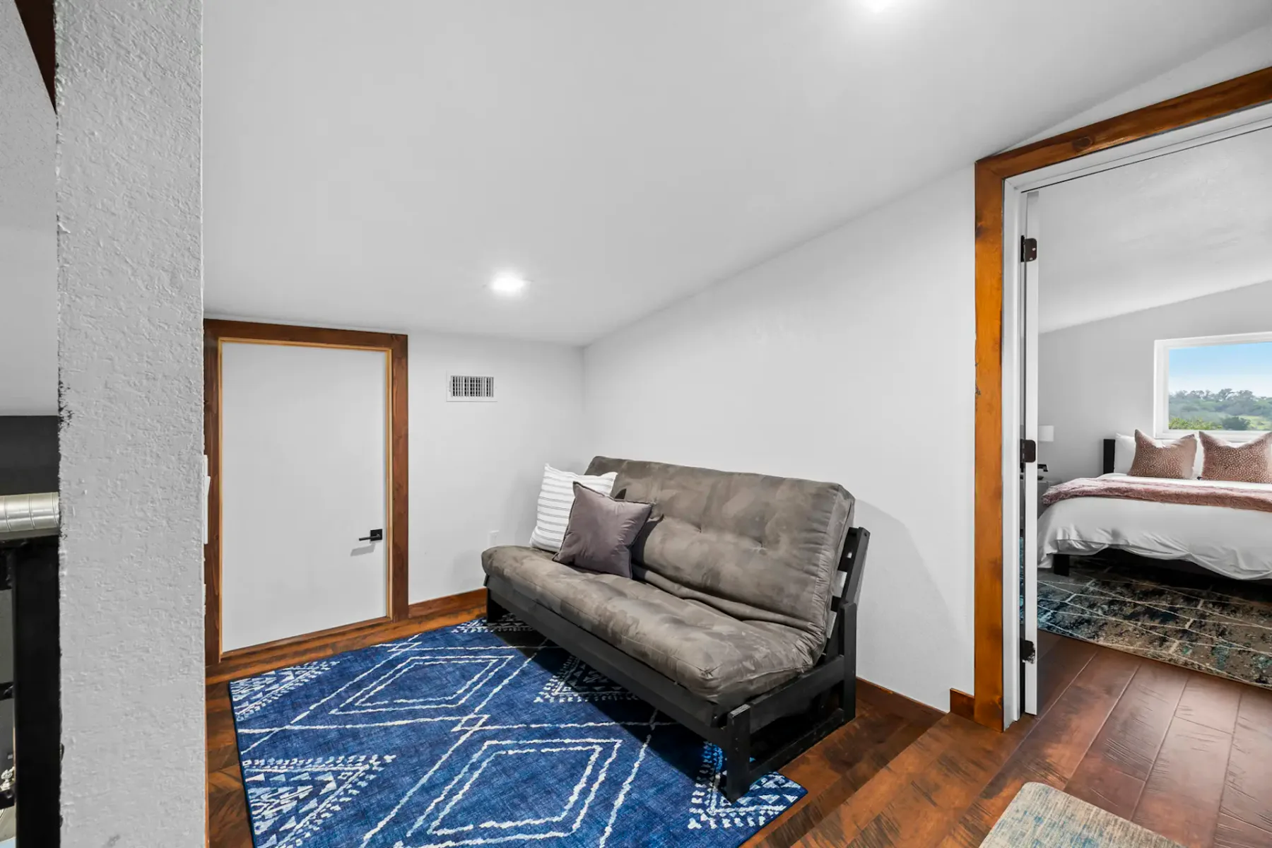 A cozy room featuring a dark futon with pillows, a blue patterned rug, and wooden flooring. An open door leads to a bedroom with a bed.