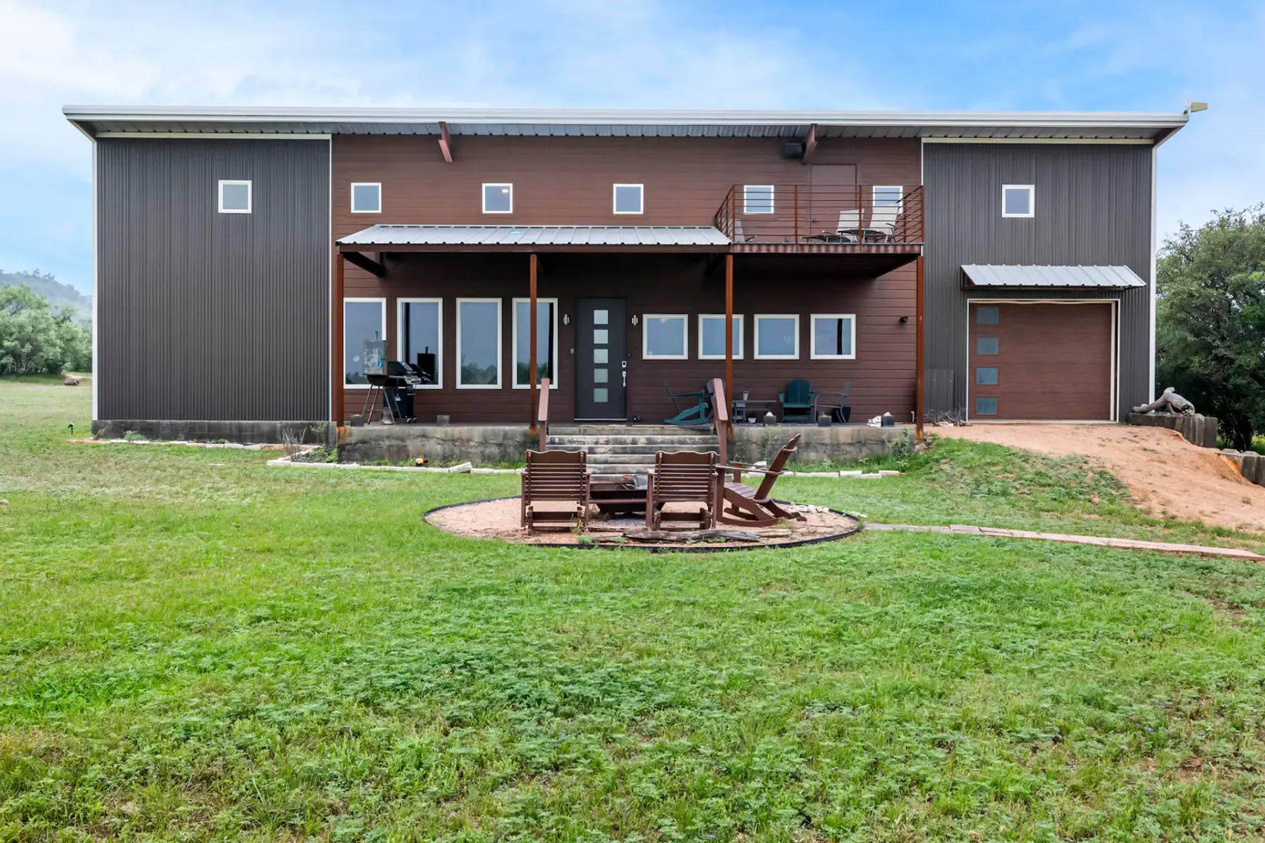 A modern two-story building with wooden and metal siding, featuring a porch, balcony, and a small fire pit area with chairs on a grassy lawn.
