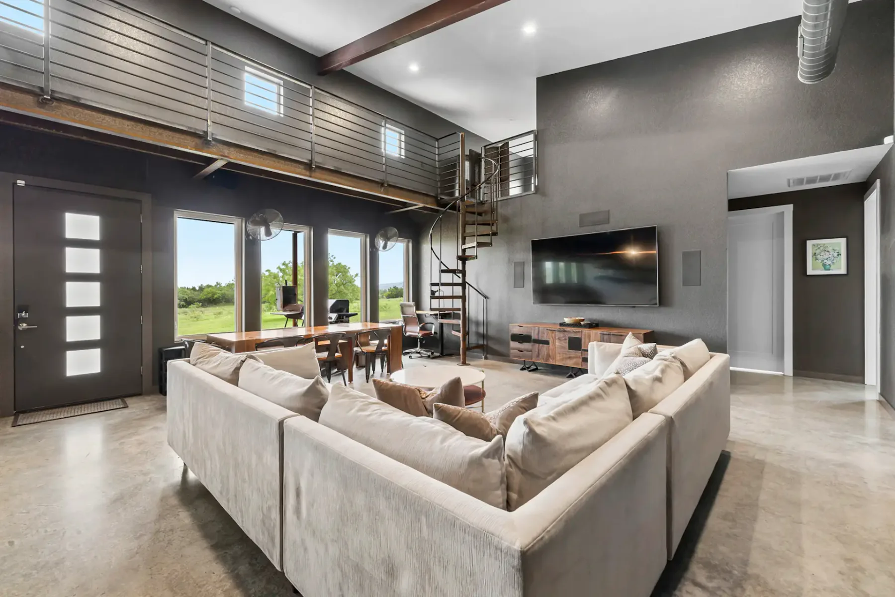 Spacious modern living room with a large beige sectional sofa, flat-screen TV, spiral staircase, and large windows overlooking greenery.