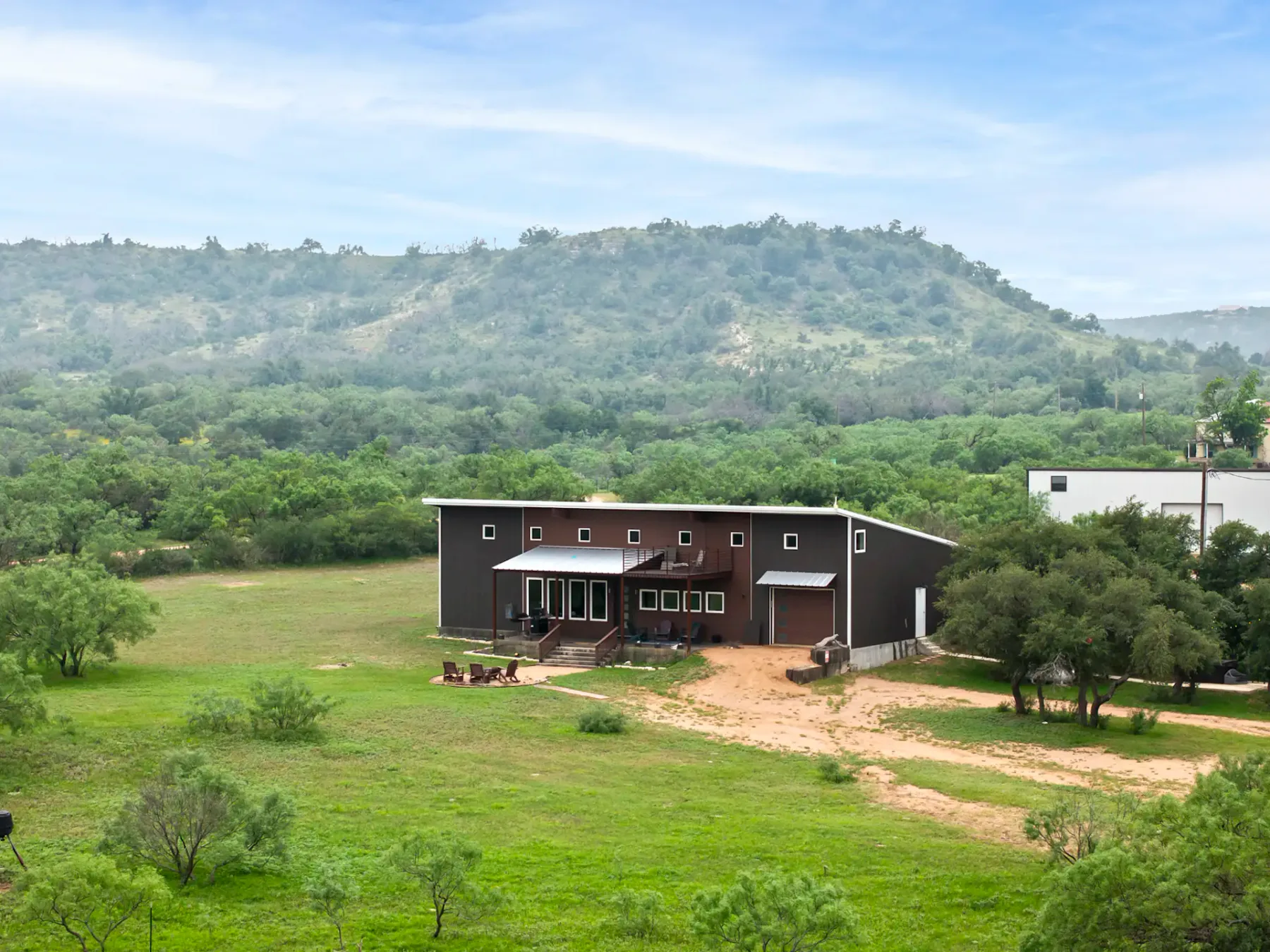 A modern two-story building with wooden and metal siding, featuring a porch, balcony, and a small fire pit area with chairs on a grassy lawn.