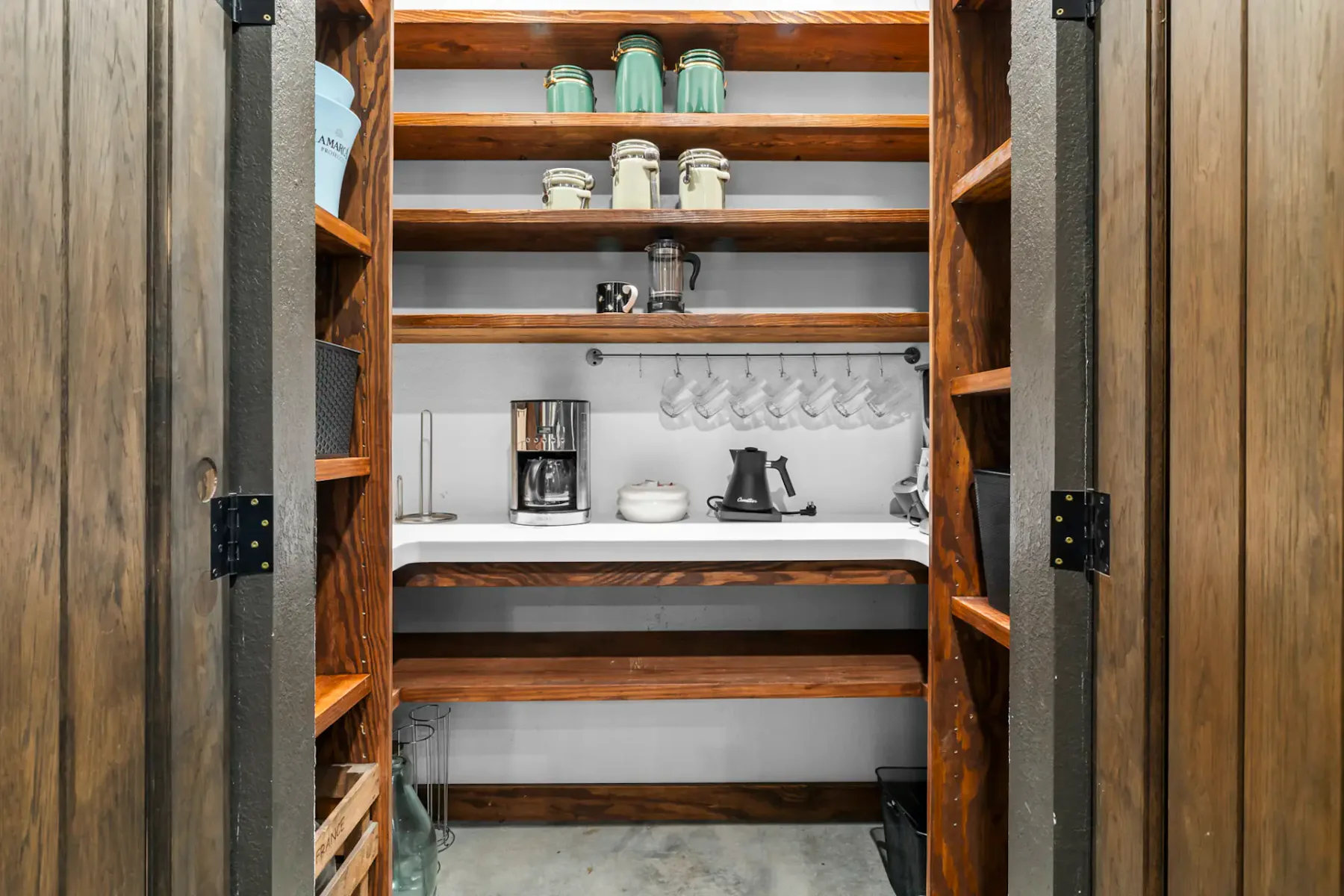 Wooden pantry with shelves holding jars, mugs, and appliances. A countertop features a coffee maker and kettle, with cups hanging above.