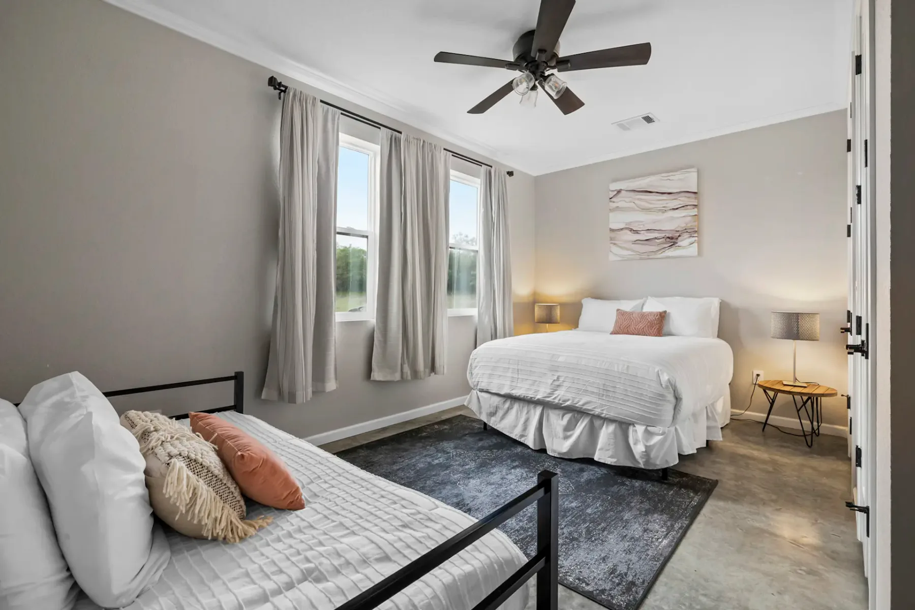 Bedroom with a double bed and a single bed, gray rug, ceiling fan, and two windows with curtains. Wall art above the bed. Neutral tones.