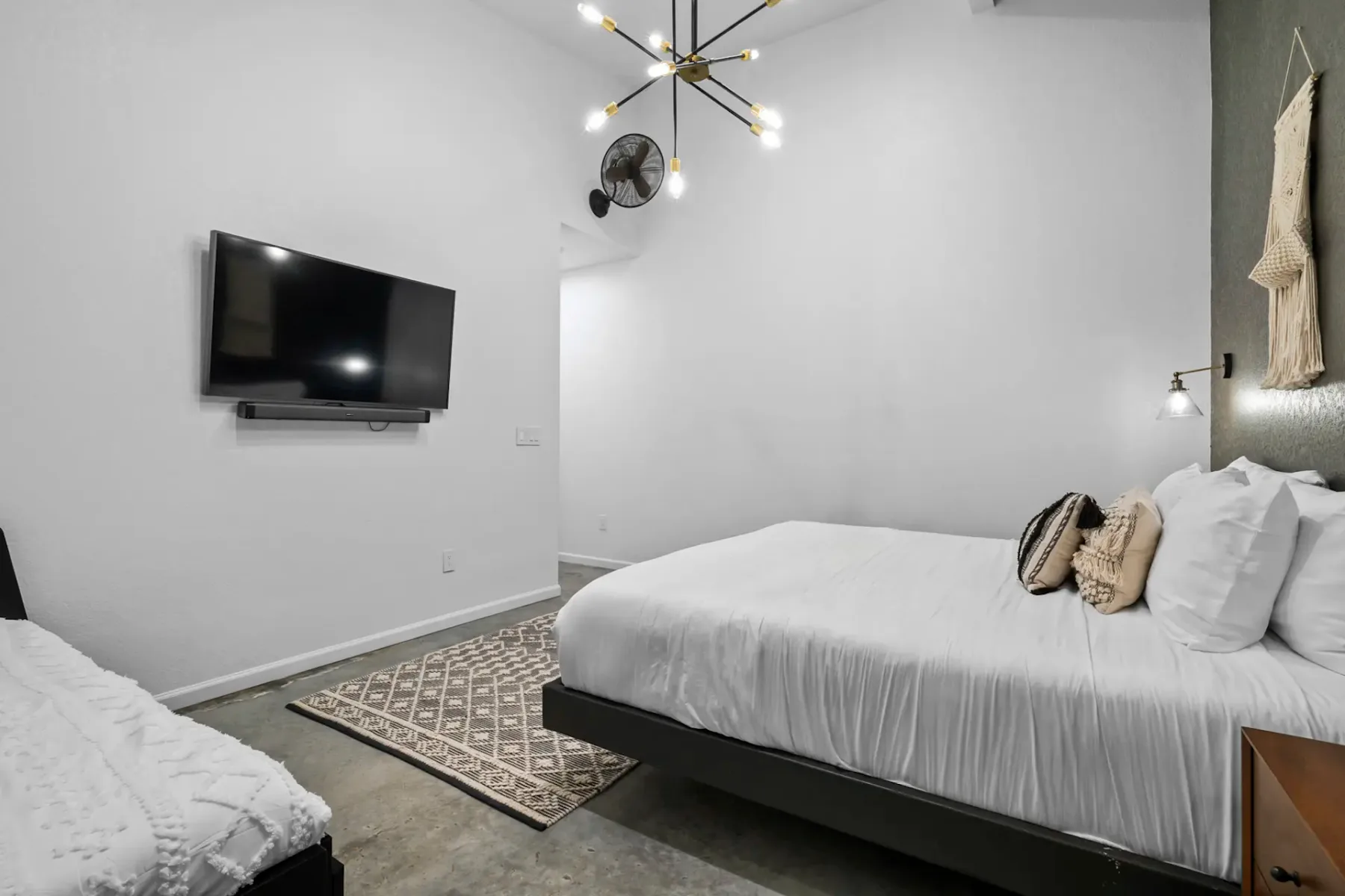 Modern bedroom with a wall-mounted TV, chandelier, double bed with white bedding, decorative pillows, and a patterned rug on a concrete floor.