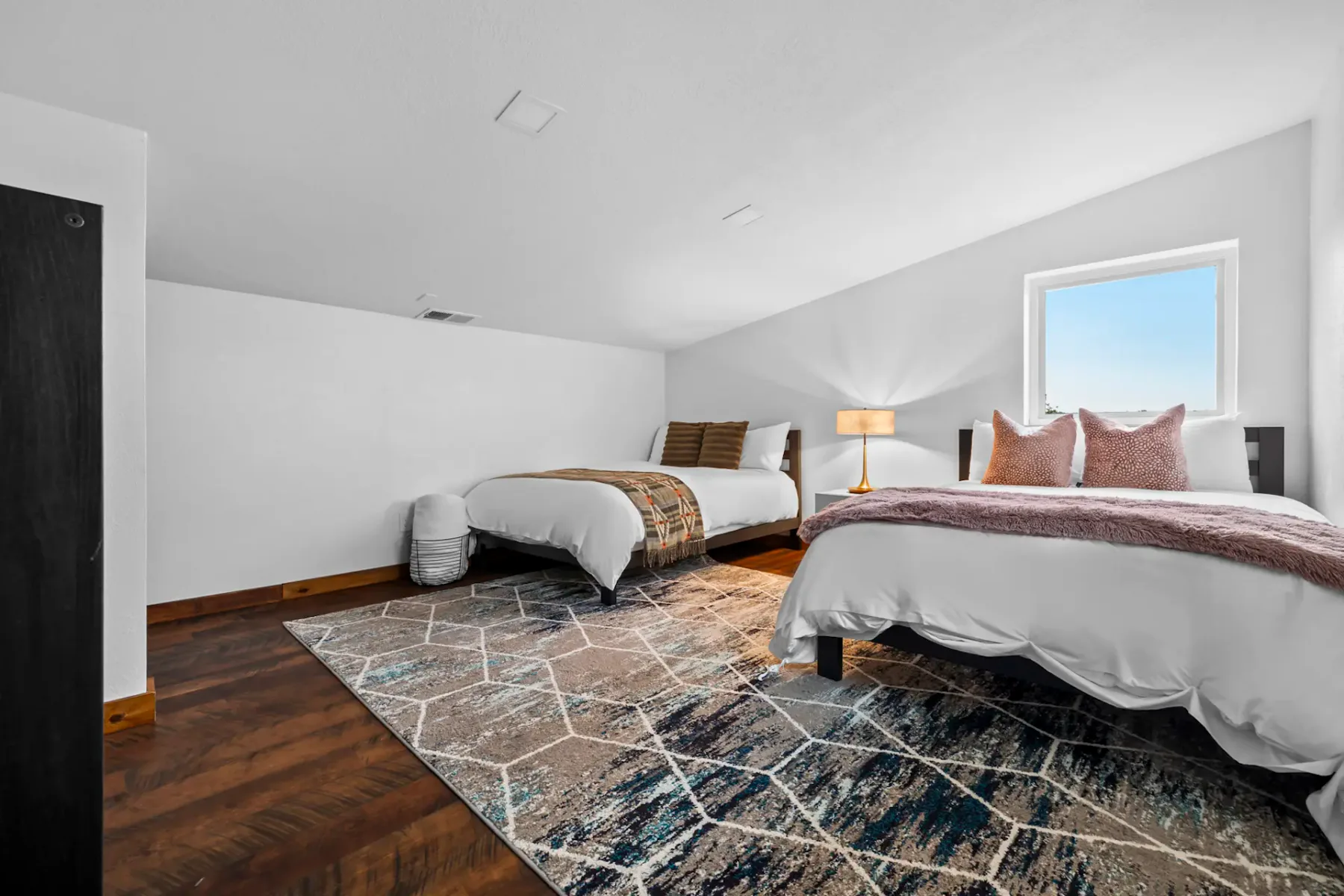 Two neatly made beds with patterned blankets in a bright room, featuring a geometric rug and two small windows letting in natural light.