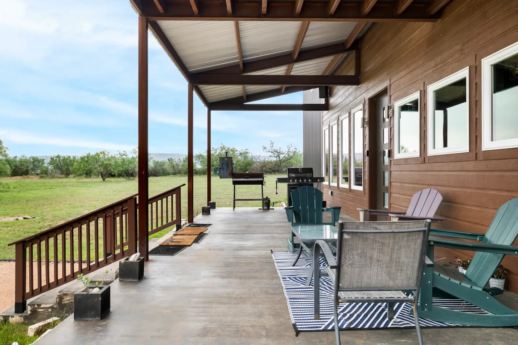 A wooden porch with a covered roof, outdoor seating, a striped rug, and a grill. It overlooks a grassy yard under a partly cloudy sky.