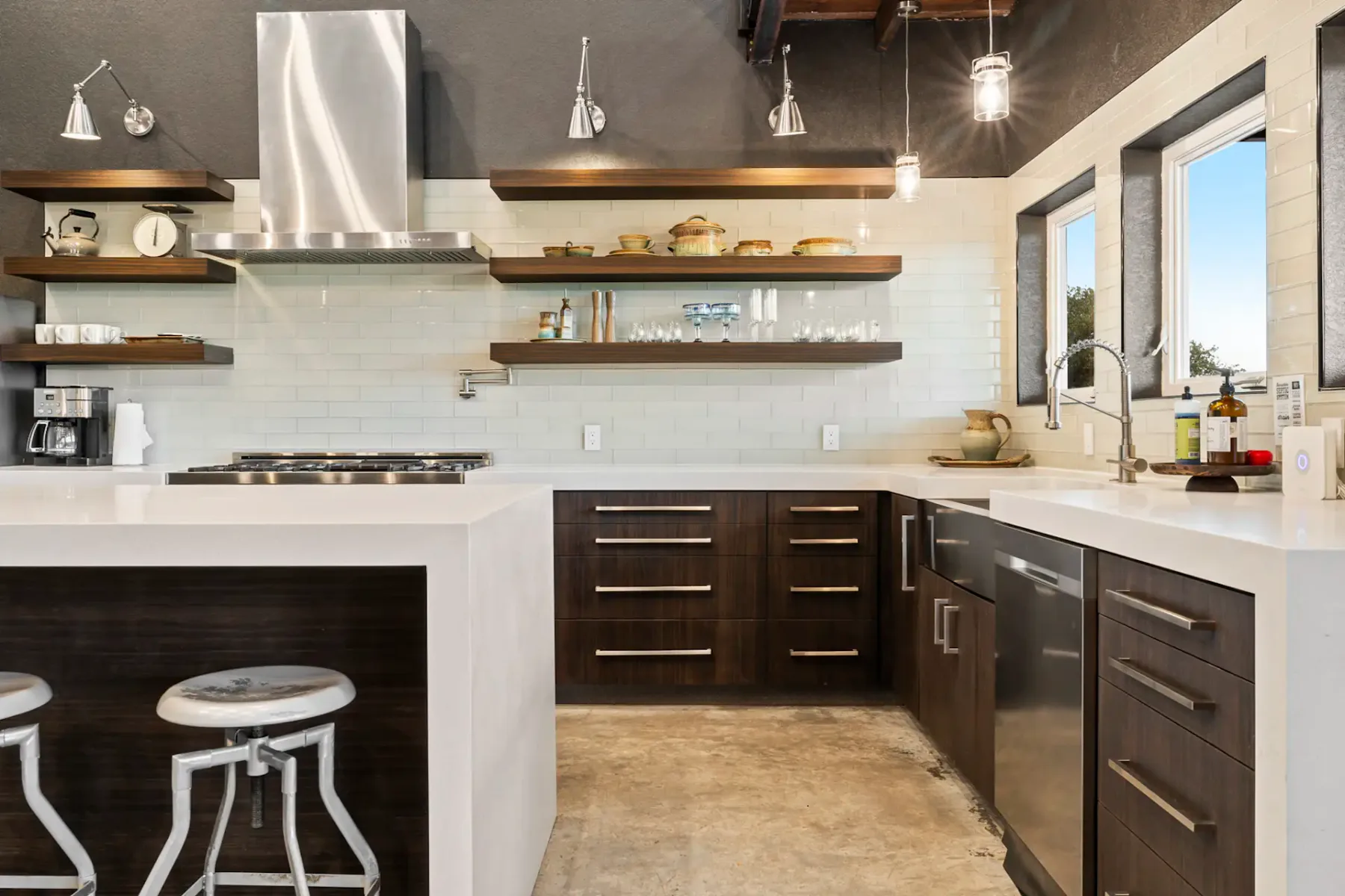 Modern kitchen with dark wood cabinets, white countertops, open shelving, hanging lights, and stainless steel appliances. Three windows bring in light.