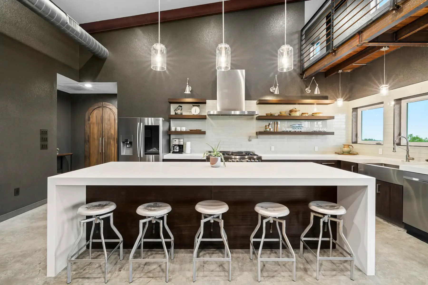 Modern kitchen with high ceiling, central white island with stools, stainless steel appliances, and open wooden shelves.