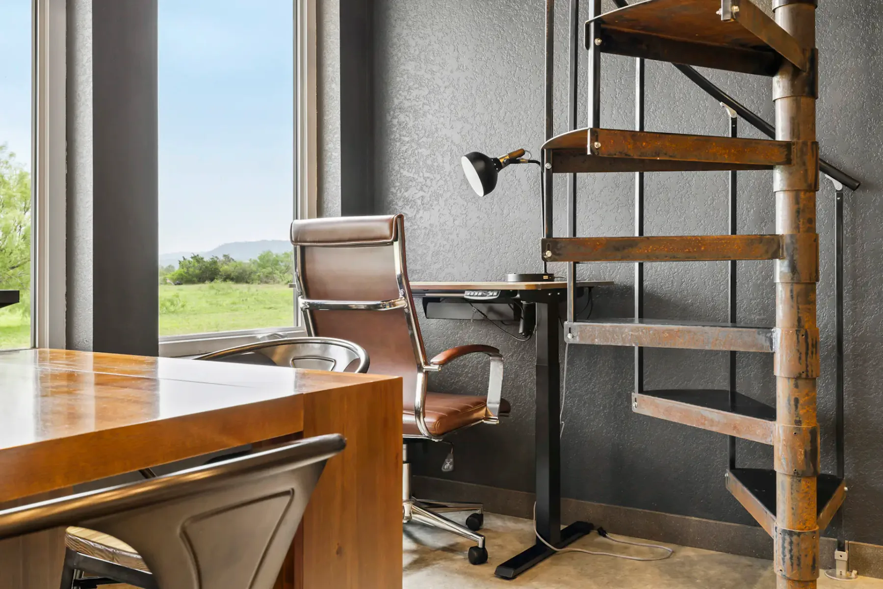 A workspace with a desk and chair beside large windows and a spiral staircase.