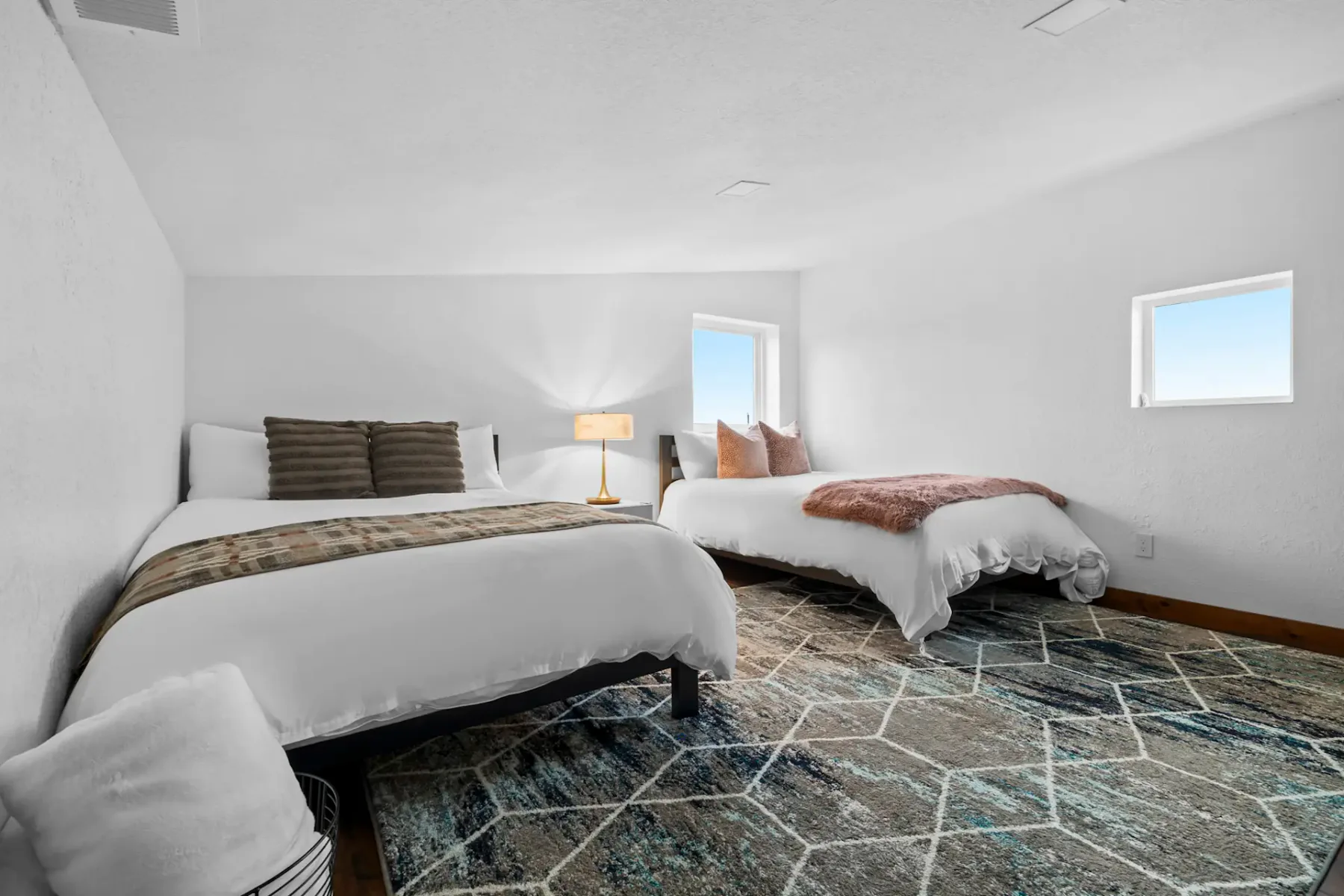 Two neatly made beds with patterned blankets in a bright room, featuring a geometric rug and two small windows letting in natural light.
