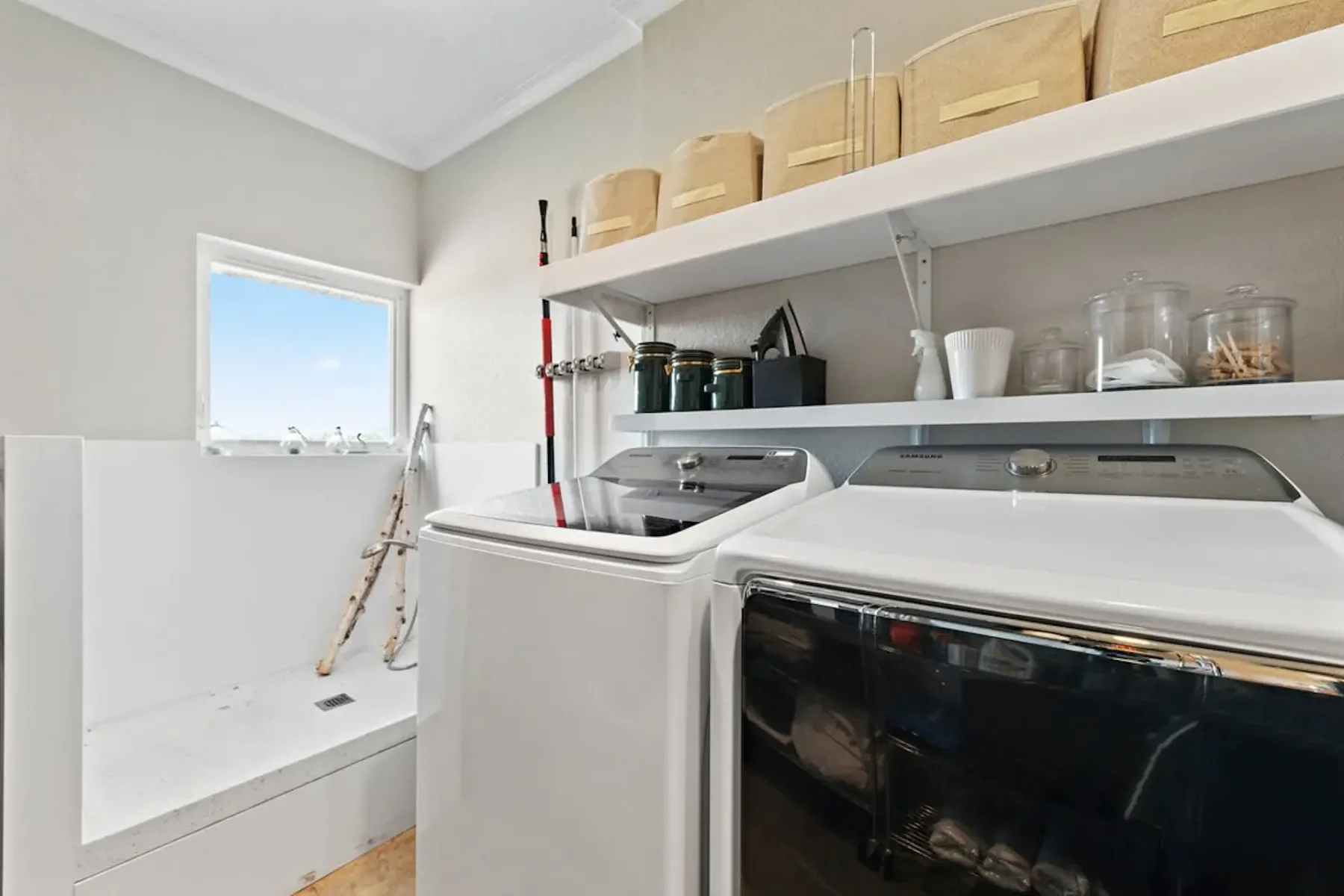 Laundry room with a washer, dryer, shelves, and a utility sink beneath a window.