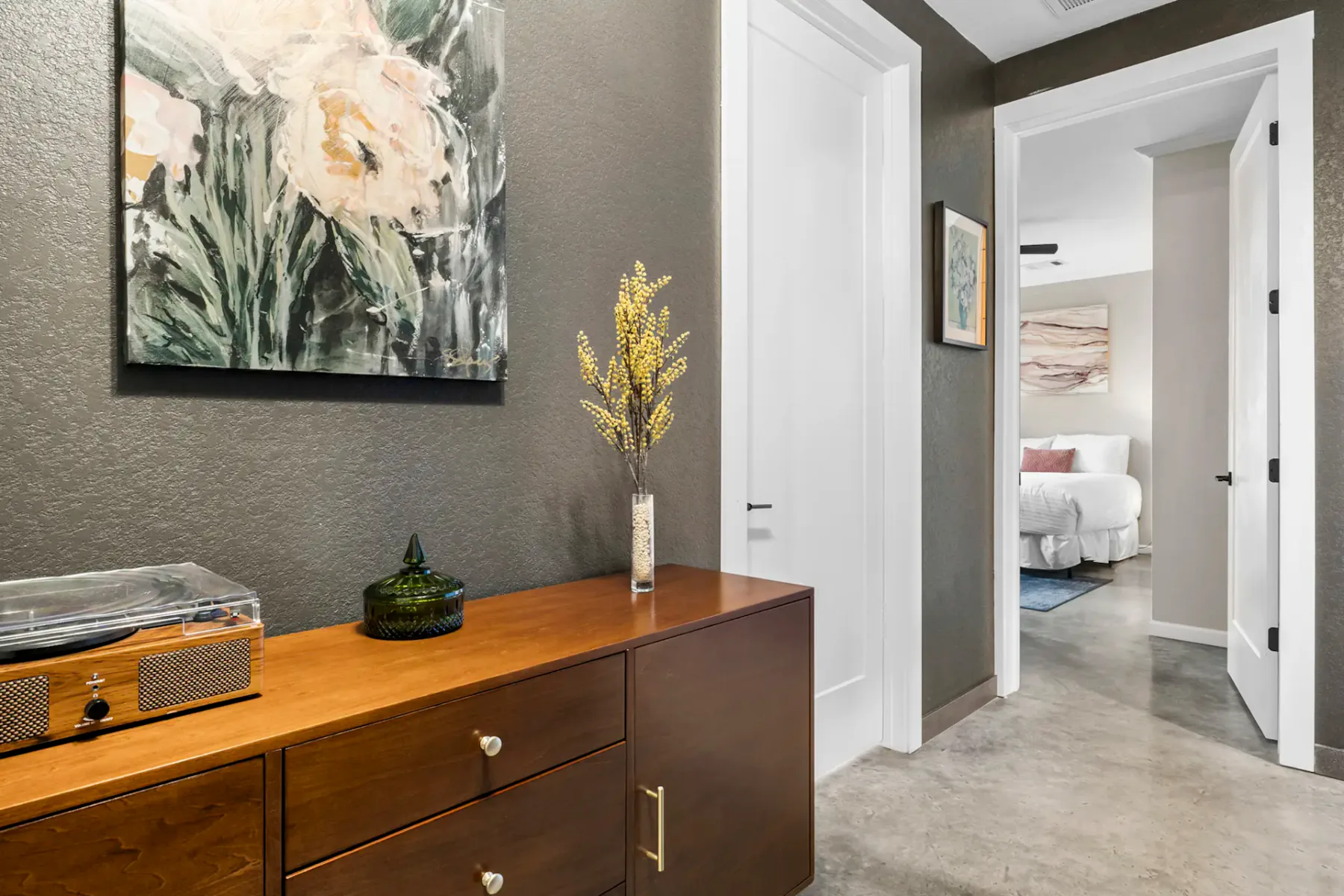 Hallway with a wooden sideboard, floral painting, and vase on top. Open door reveals a bedroom with a bed and decorative art on the walls.