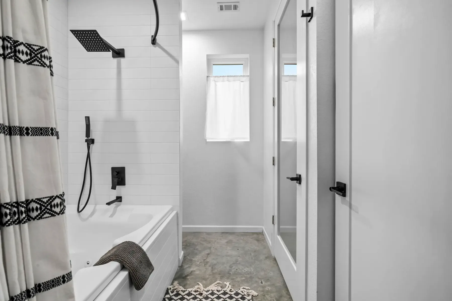 Modern bathroom with a white tub, black fixtures, and geometric shower curtain. Concrete floor and glass-paneled door. Natural light from a window.