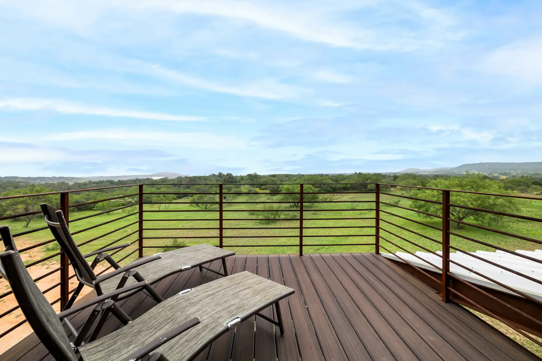 Wooden deck with two lounge chairs, overlooking a lush green landscape with a clear blue sky.