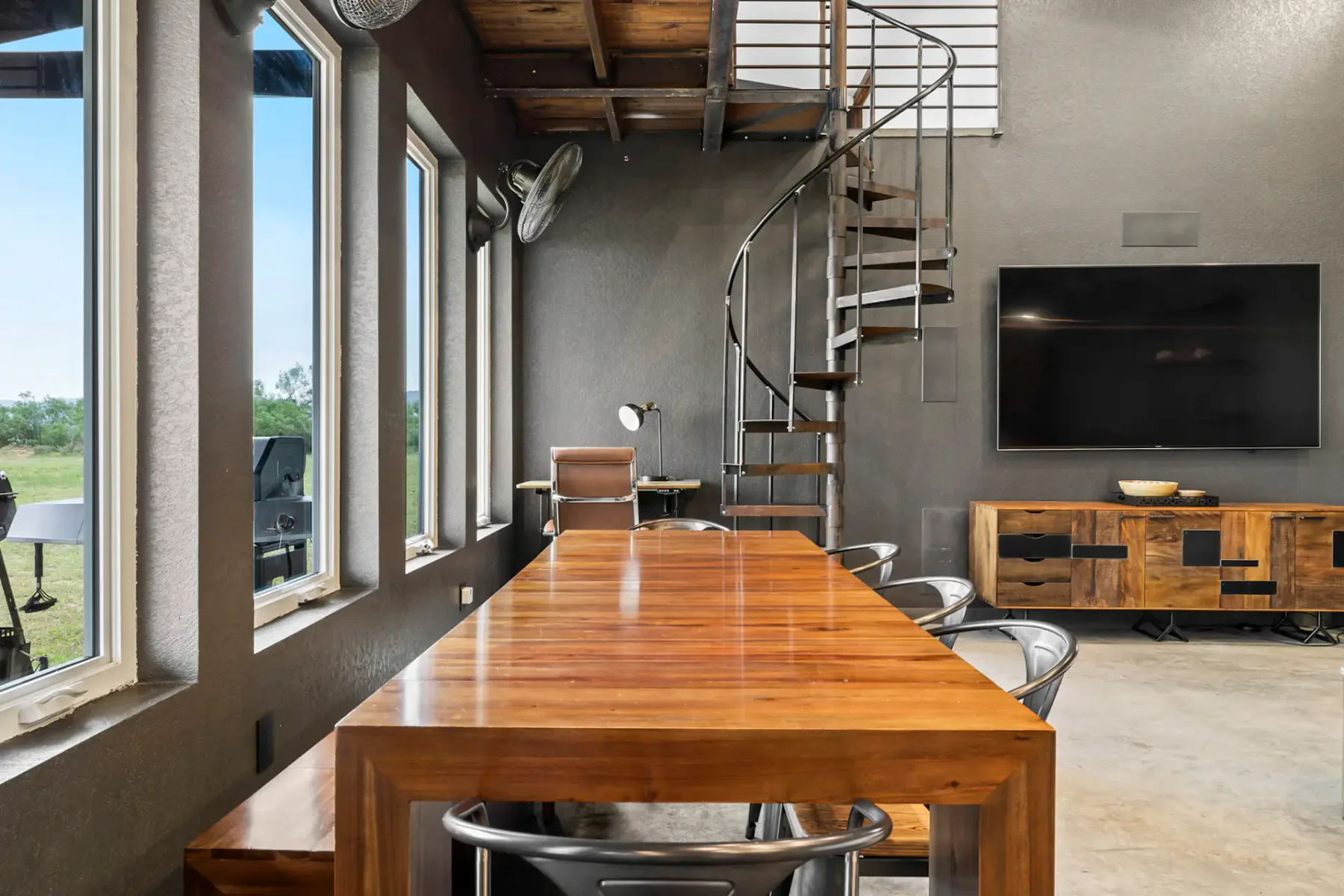 Modern room with a long wooden table, metal chairs, spiral staircase, large TV, and windows offering a view of greenery outside.