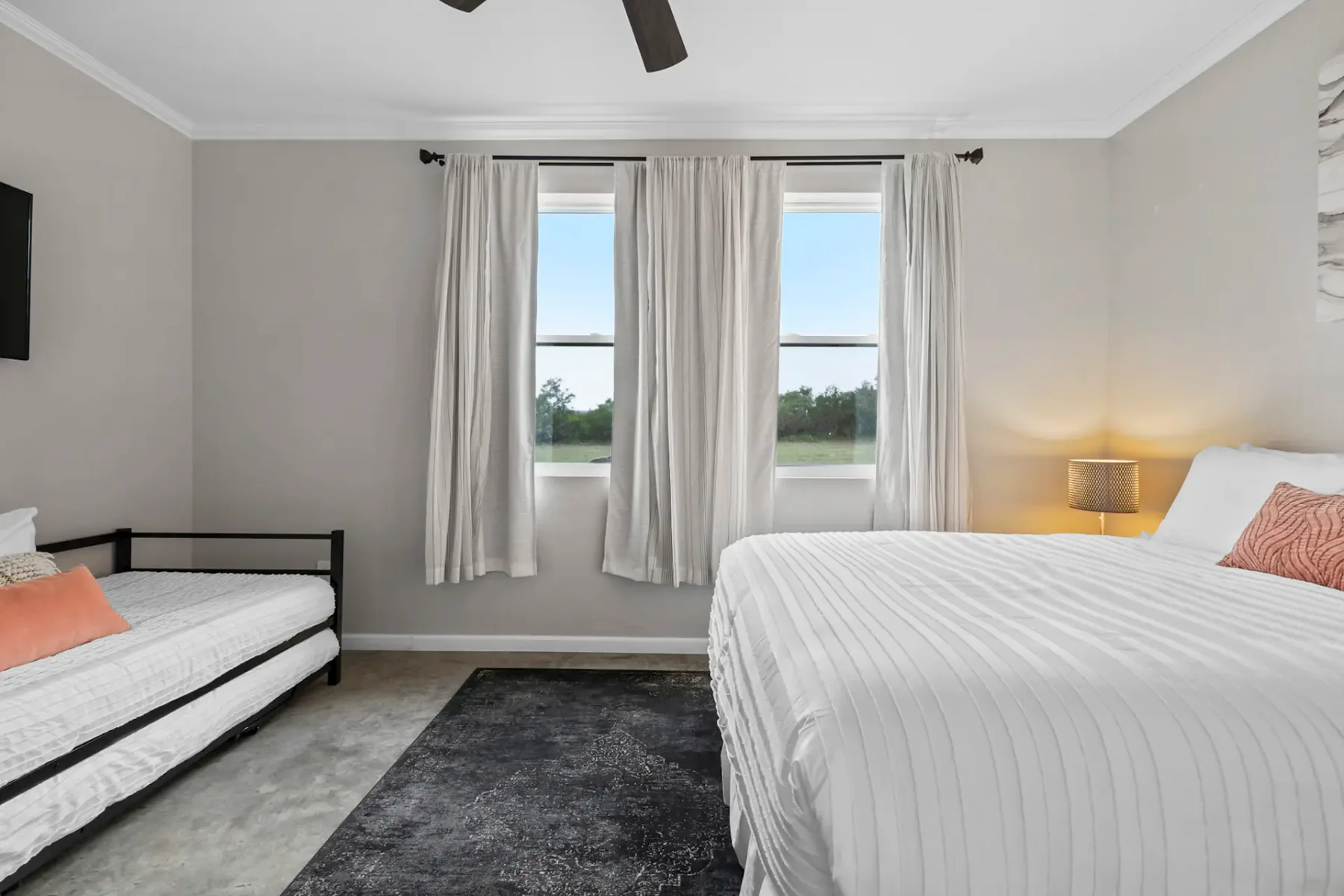 Bedroom with a double bed and a single bed, gray rug, ceiling fan, and two windows with curtains. Wall art above the bed. Neutral tones.