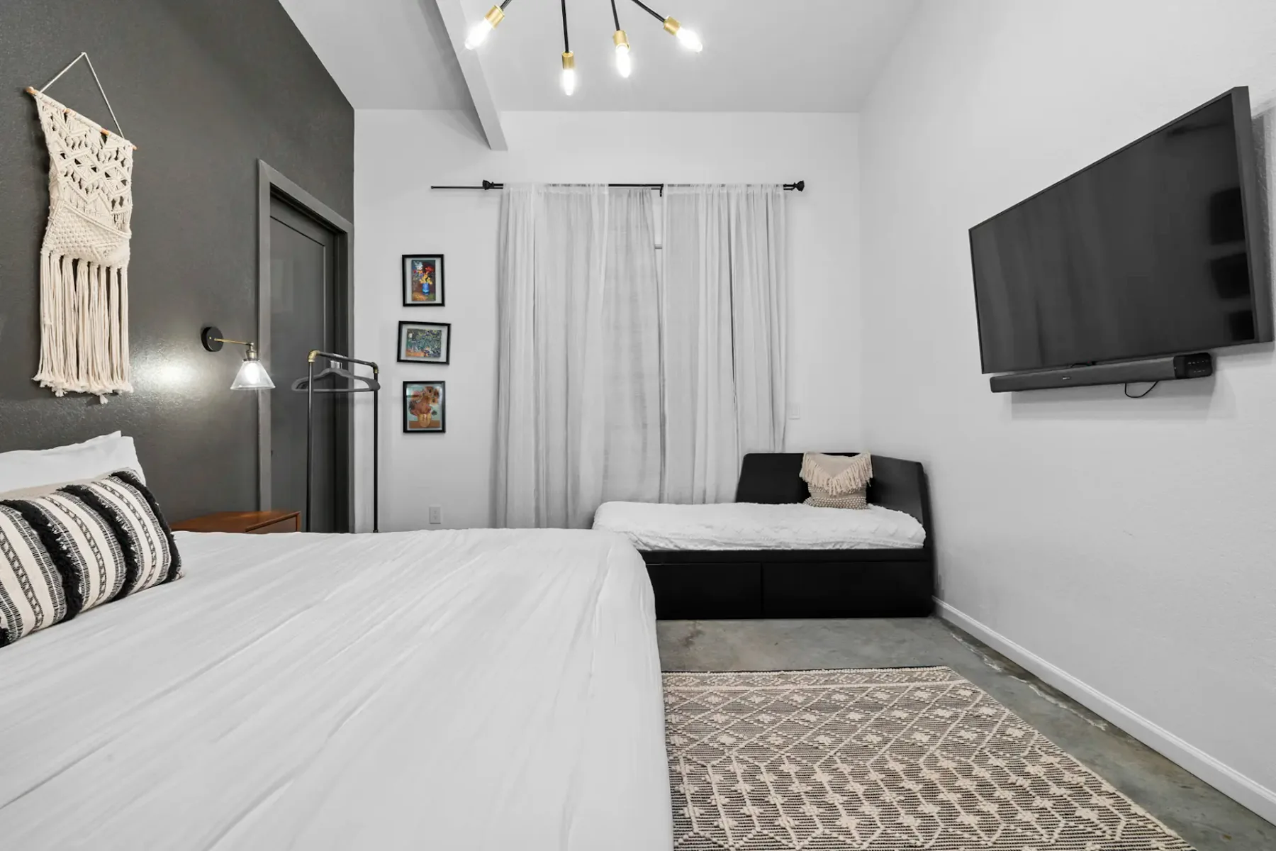 Modern bedroom with a double bed, wall-mounted TV, sofa, wall tapestry, and framed art. Neutral tones with a touch of bohemian decor.