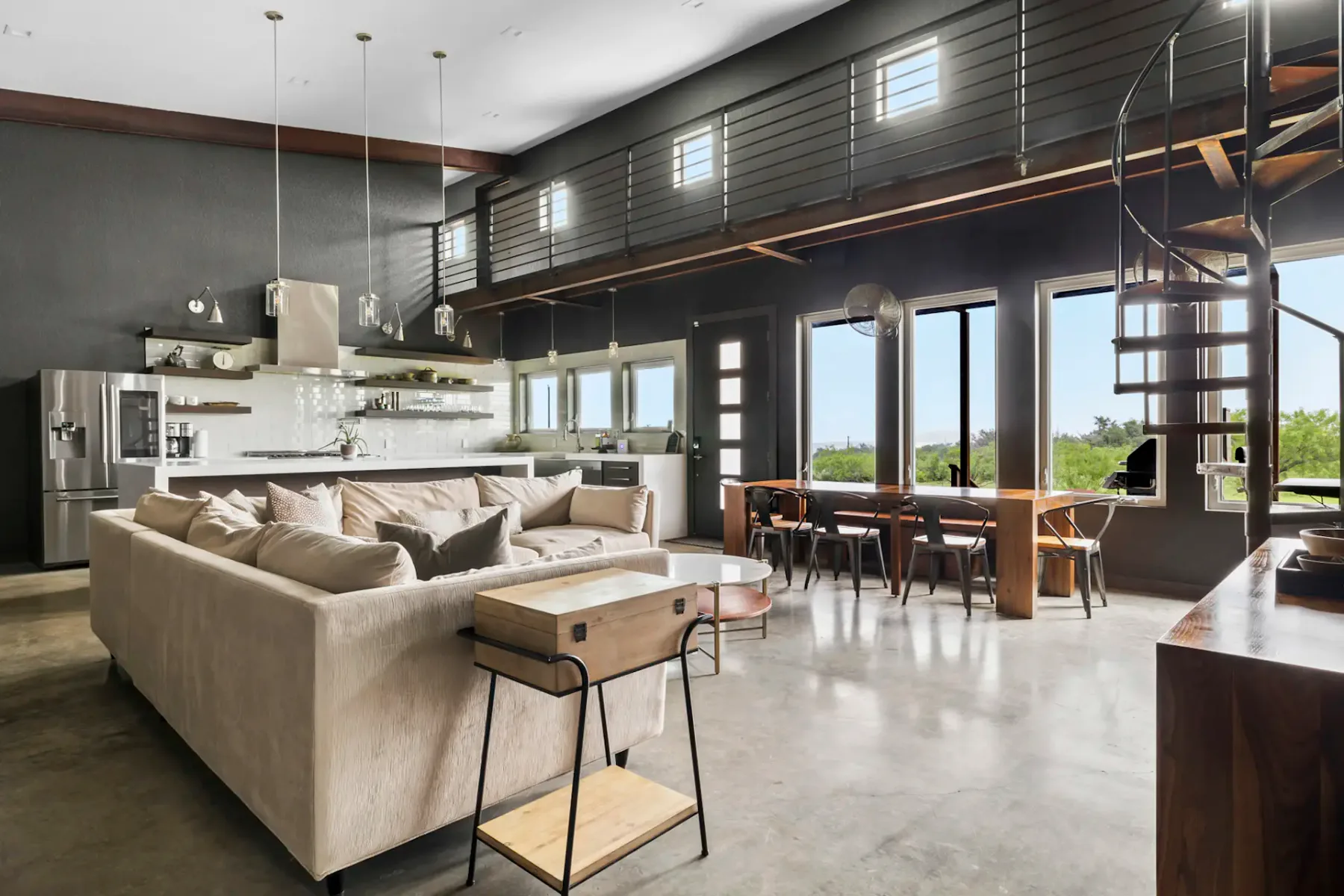 Modern open-concept living space with a sectional sofa, dining table, sleek kitchen, spiral staircase, and large windows with outdoor views.