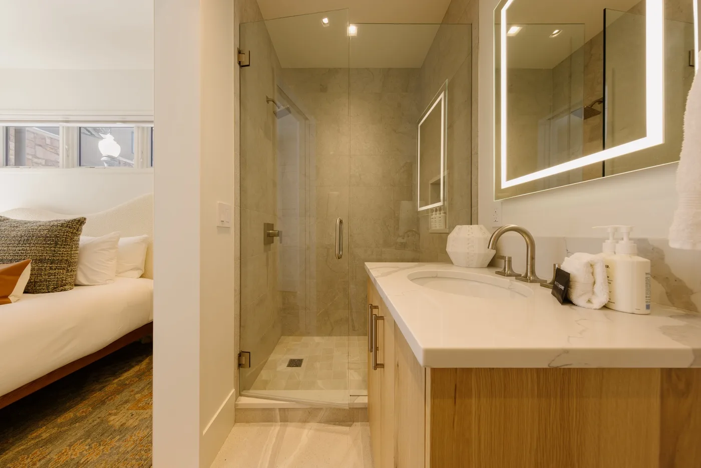 Un lavabo de baño está junto a una ducha con mampara de vidrio; se ve un dormitorio por la puerta.