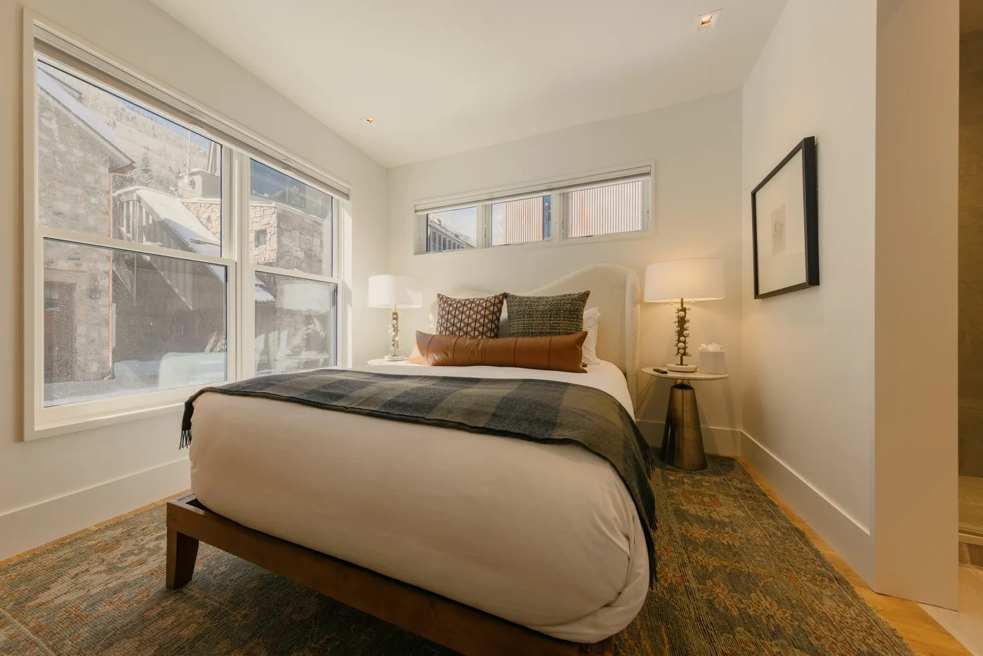 Un dormitorio con una cama bajo ventanas grandes y un cuadro en la pared.