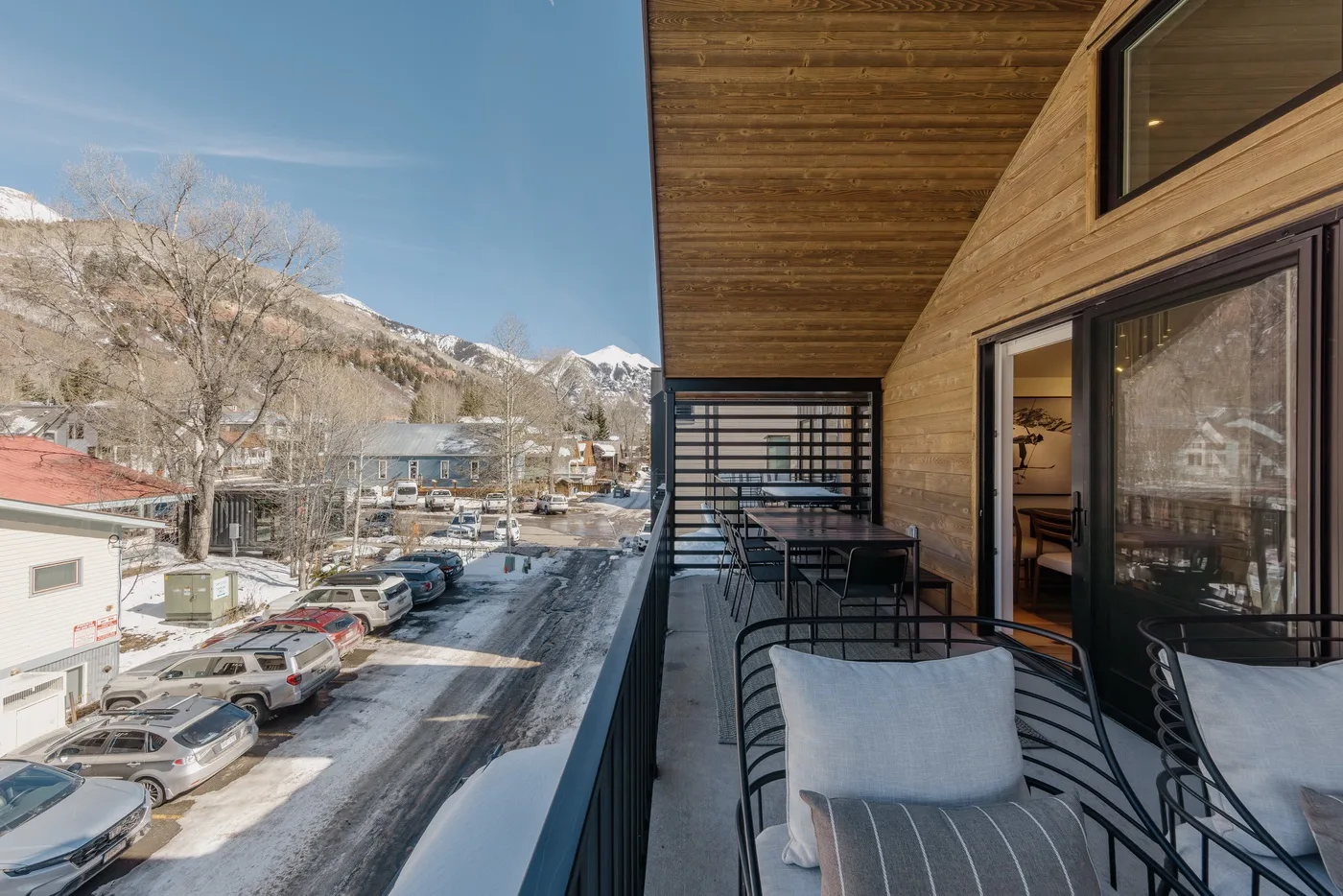 A balcony with outdoor seating overlooks a snow-dusted street and mountains.