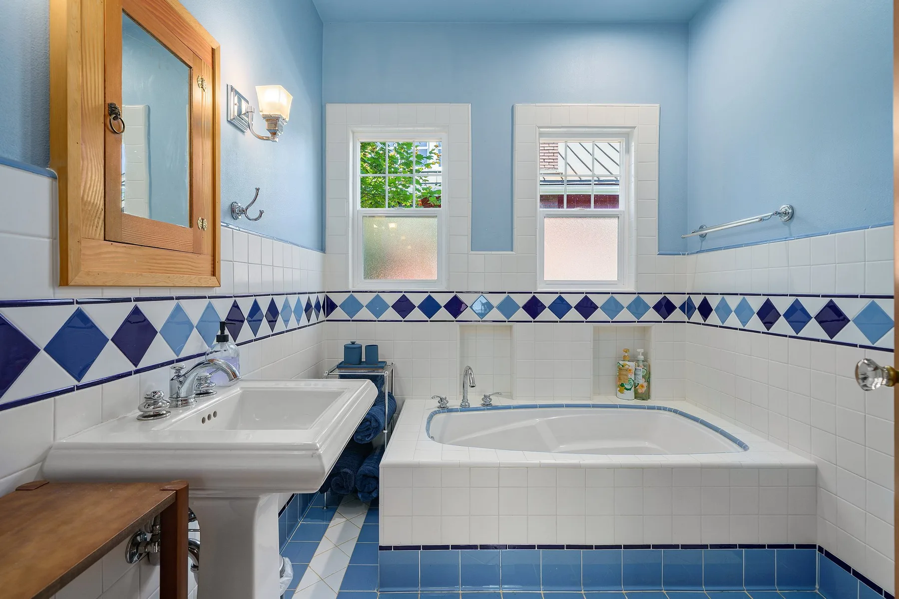 Un baño alicatado con bañera, lavabo y dos ventanas.