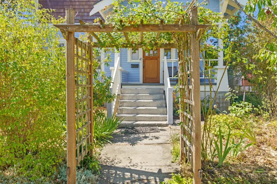 Un sendero de jardín pasa bajo una pérgola de madera hacia la entrada de una casa.