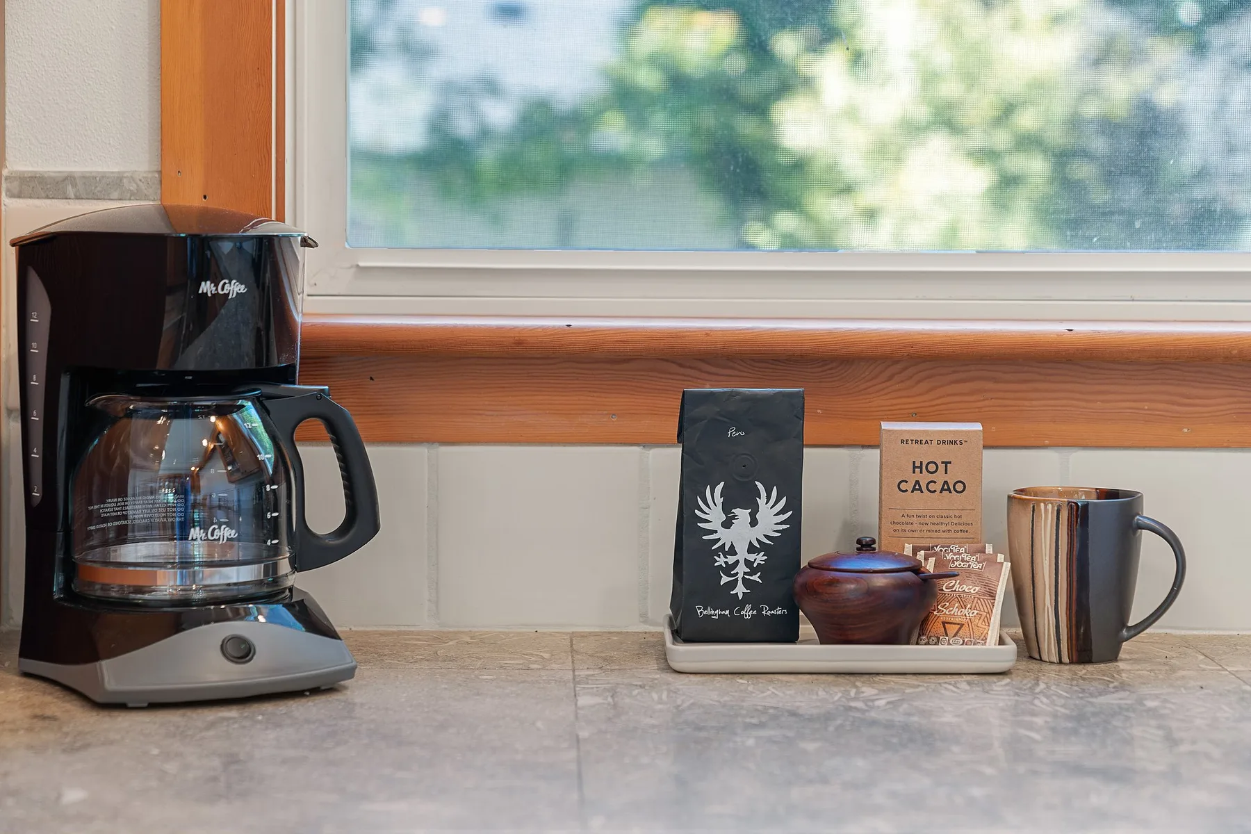 Una encimera de cocina con una cafetera, tazas y sobres de bebidas bajo una ventana.