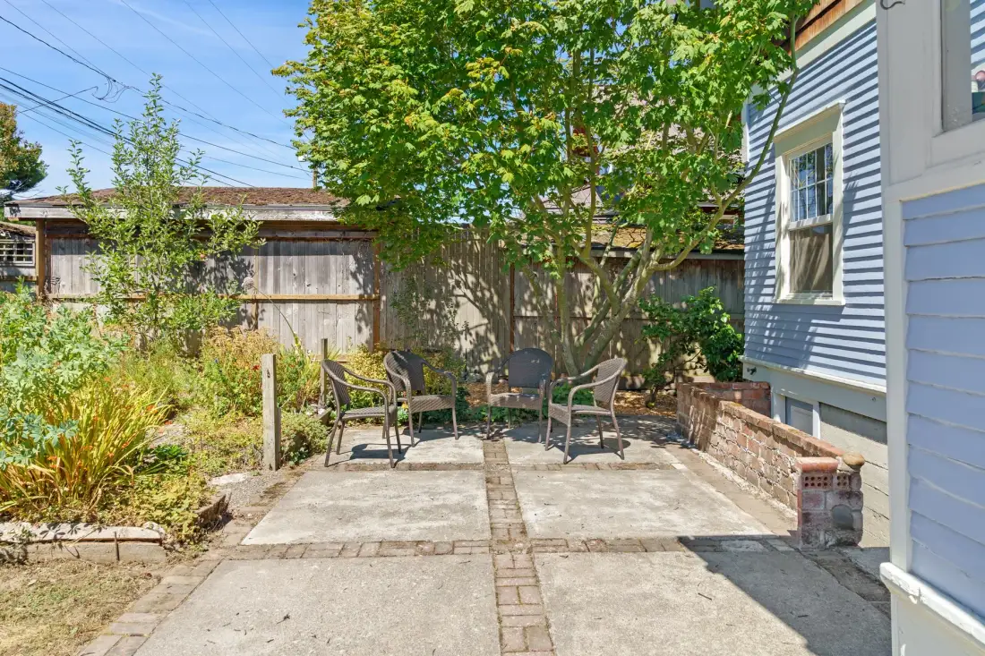 Una zona exterior pavimentada con asientos está junto a una casa bajo árboles frondosos.
