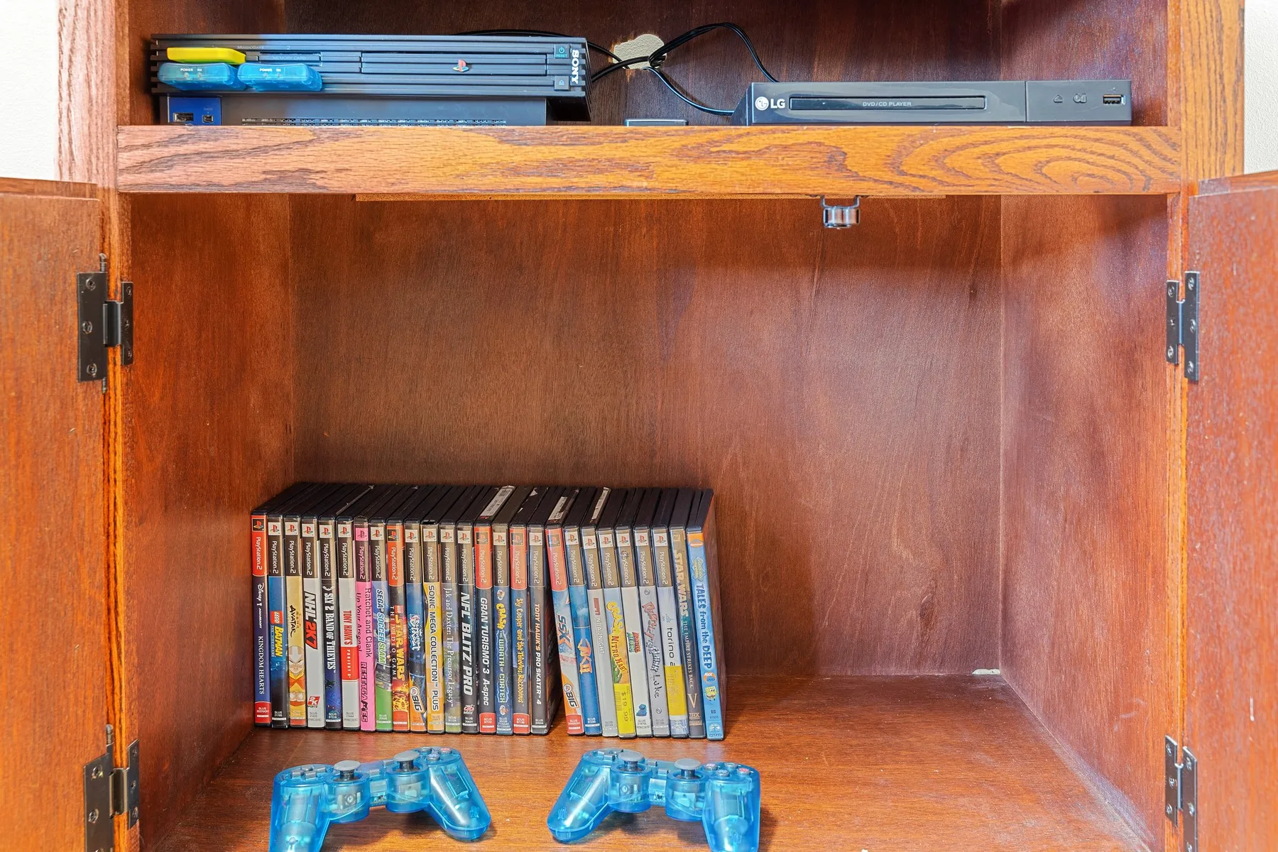 Un armario de madera abierto contiene cajas de videojuegos, mandos y dispositivos electrónicos.