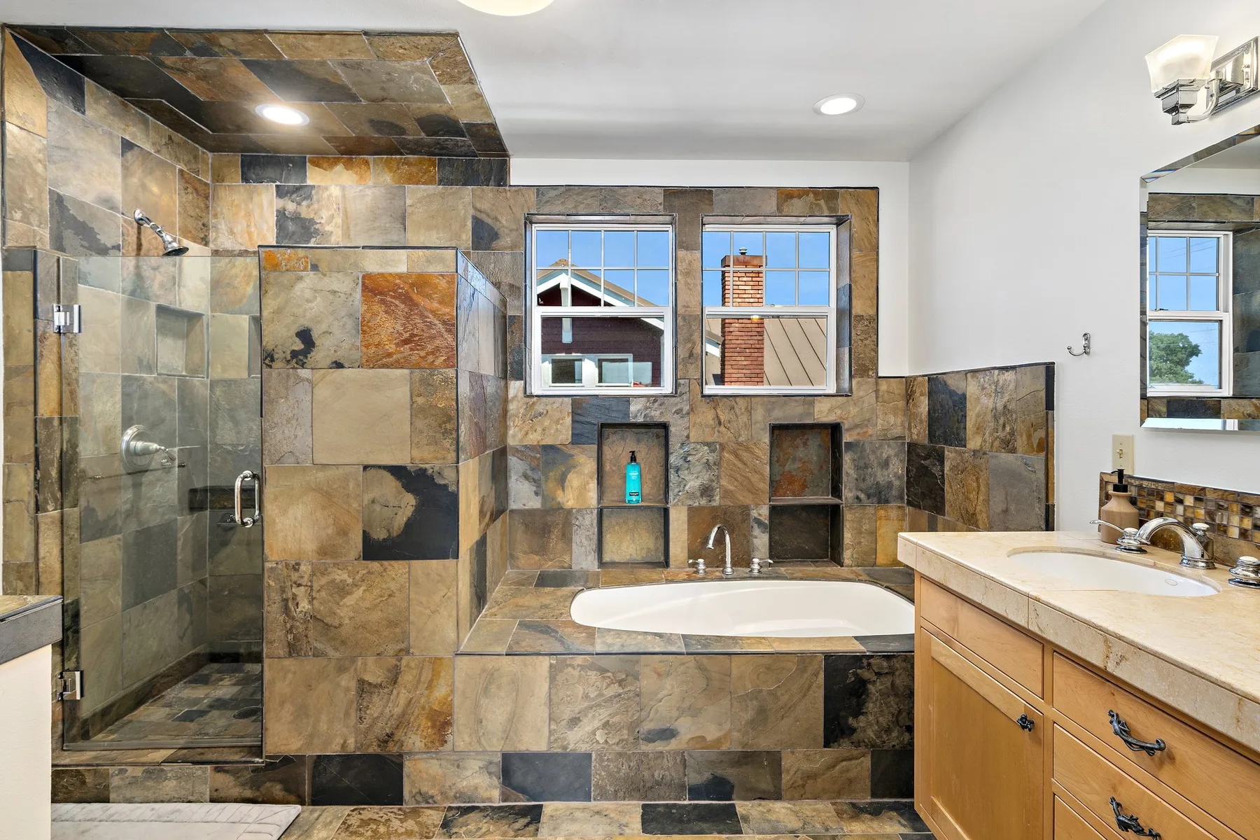 Un baño revestido de azulejos con bañera, ducha de vidrio y tocador bajo dos ventanas.