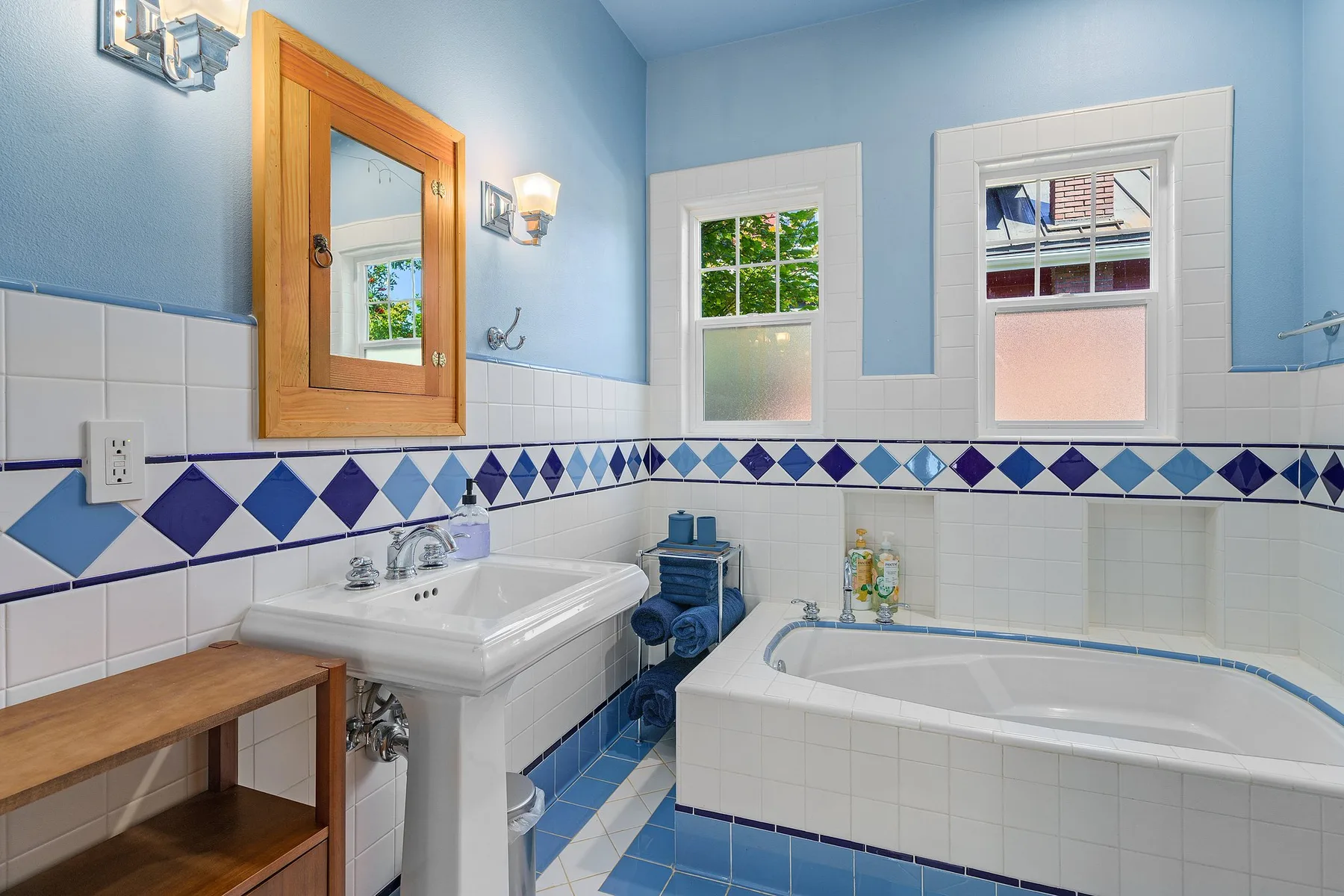 Un baño azul y blanco con bañera, lavabo y dos ventanas.