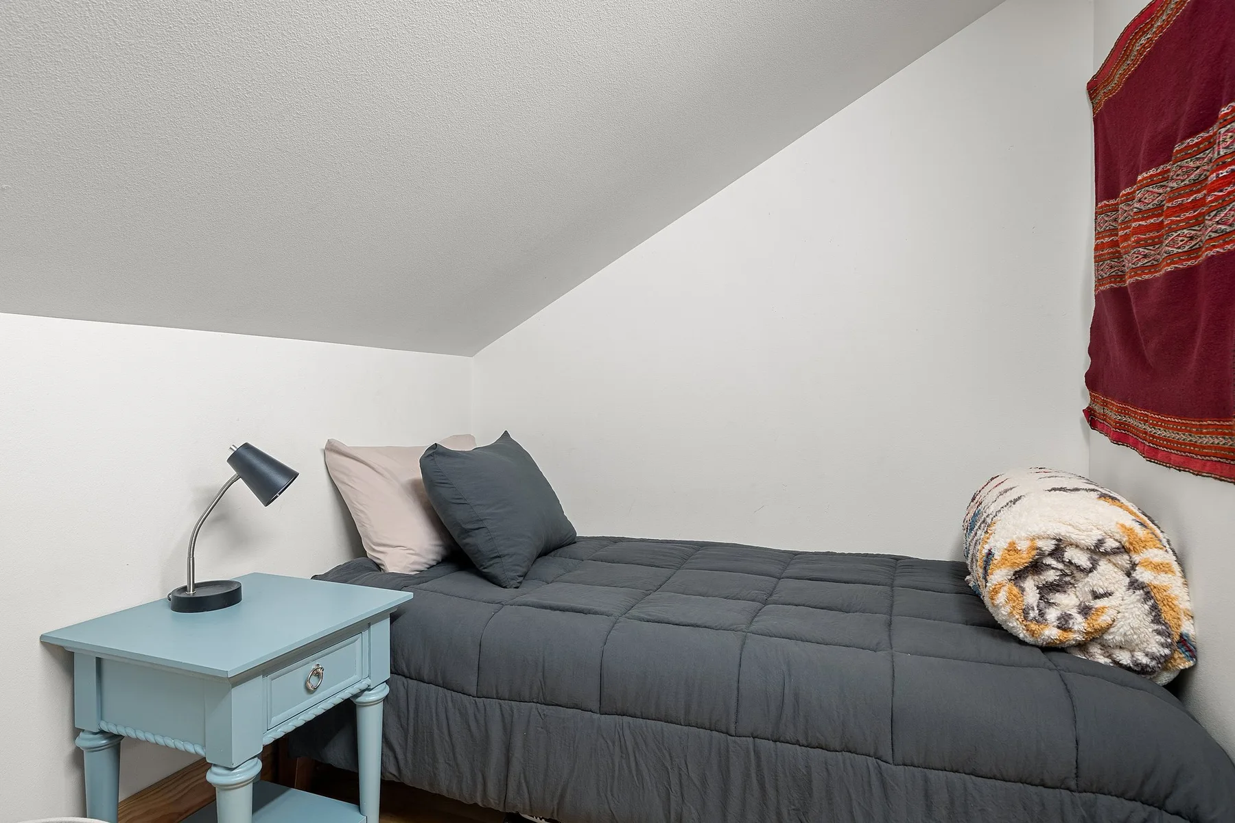 Un dormitorio con una cama gris y una pequeña mesa azul junto a la cama.