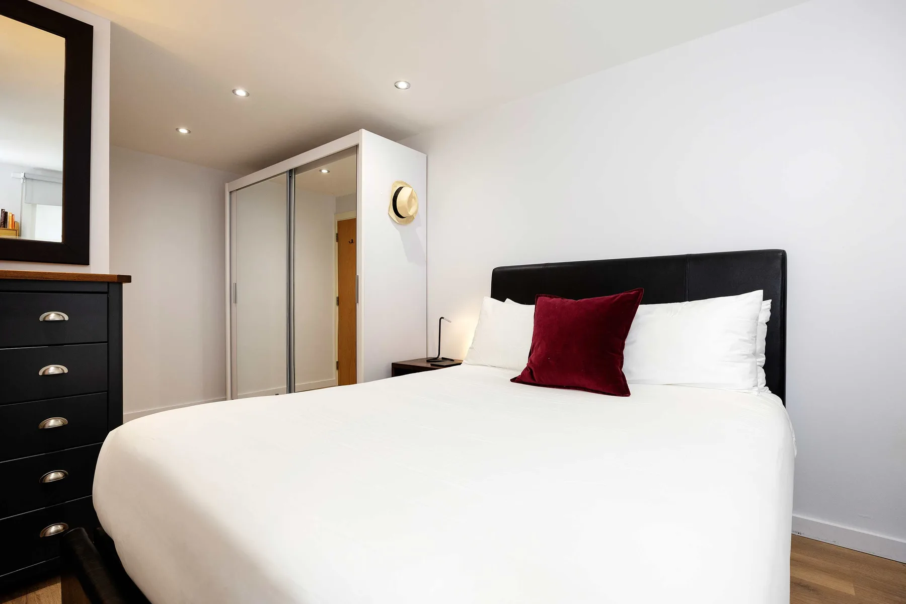 A bedroom with a white bed, dark headboard, and red accent pillow.