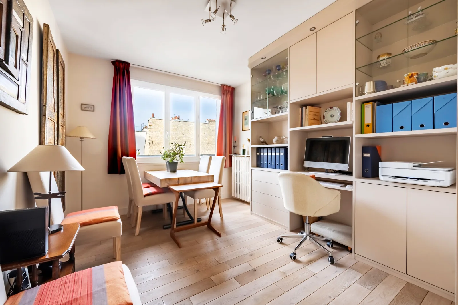 Una habitación luminosa con escritorio y estanterías junto a una mesa de comedor cerca de las ventanas.