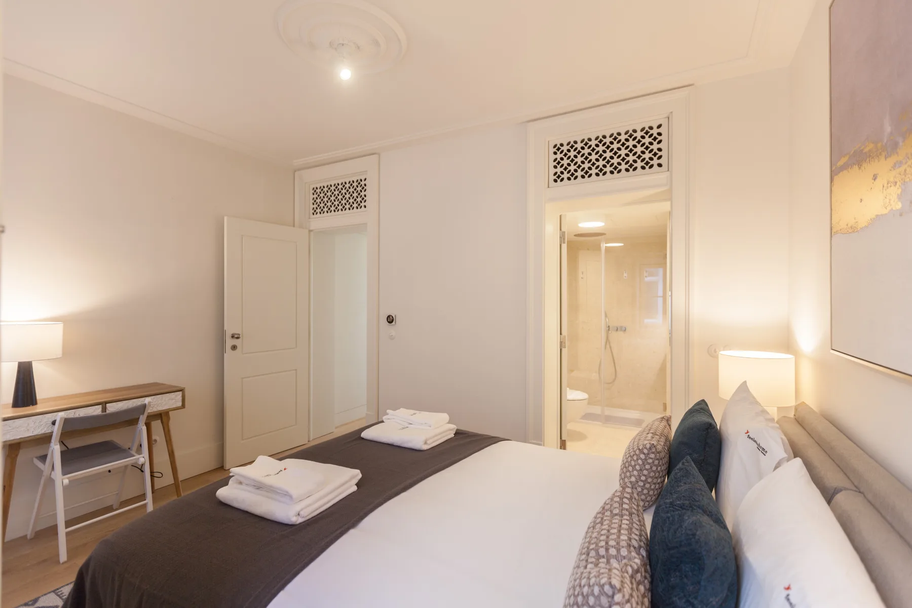 Un dormitorio con una cama bien tendida, lámparas de noche y una puerta al baño.