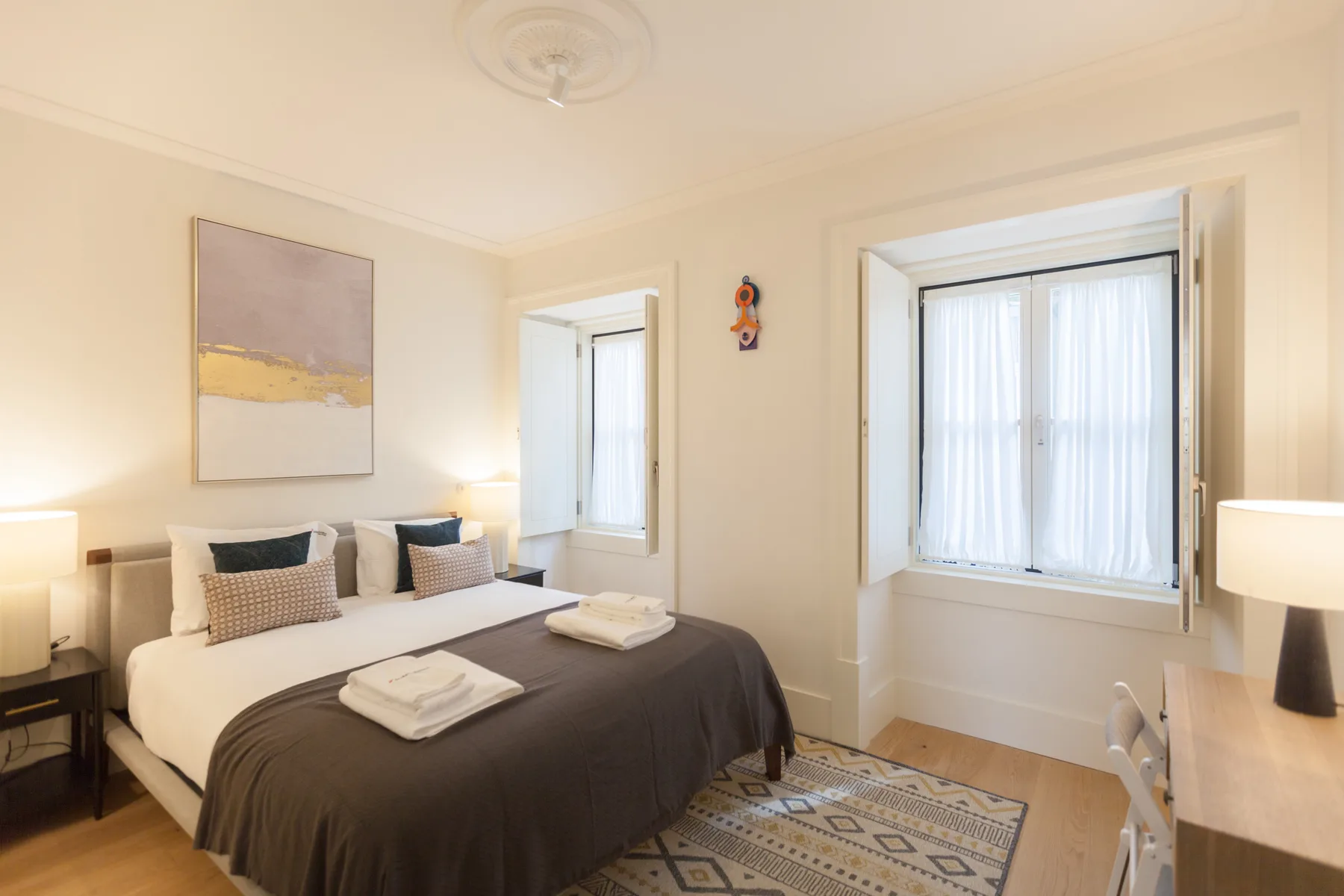 Un dormitorio luminoso con una cama bien hecha, lámparas de noche y ventanas con cortinas.