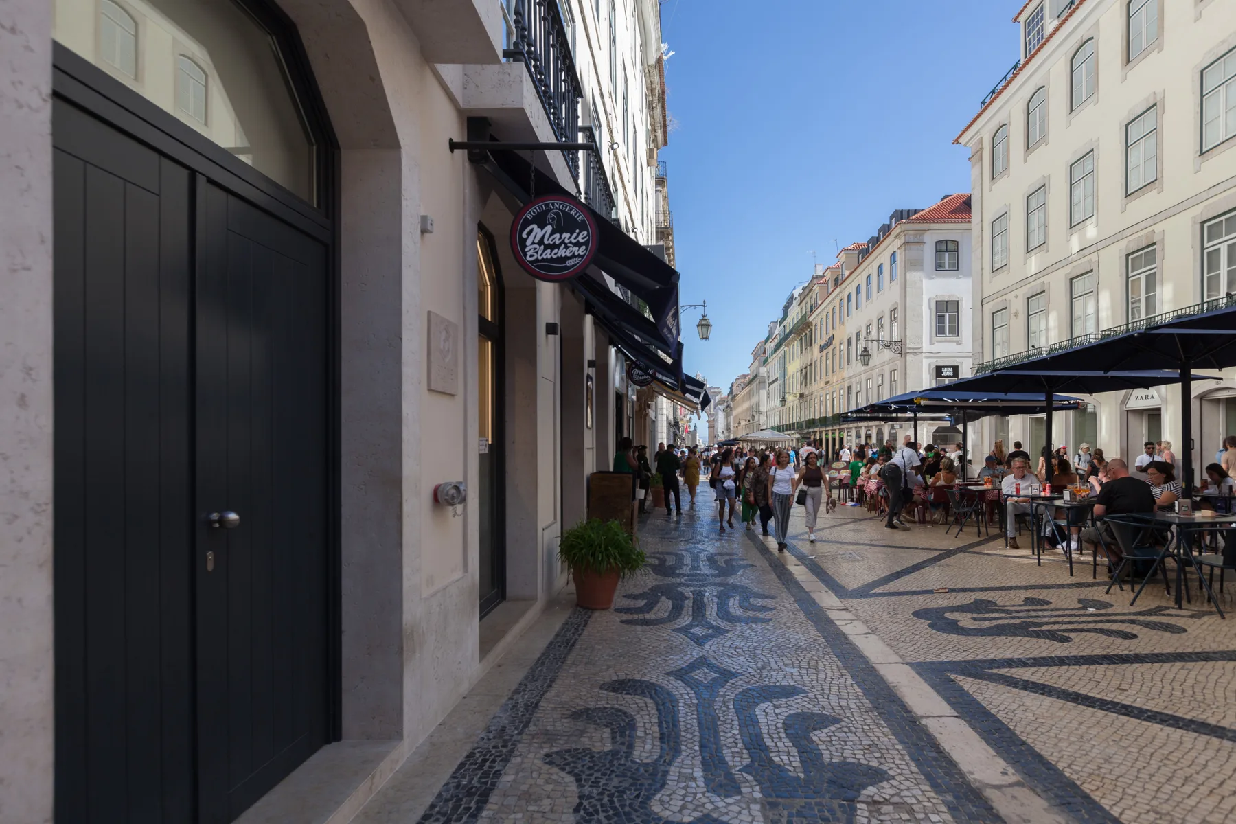 Una calle peatonal con edificios, mesas de café al aire libre y pavimento estampado.