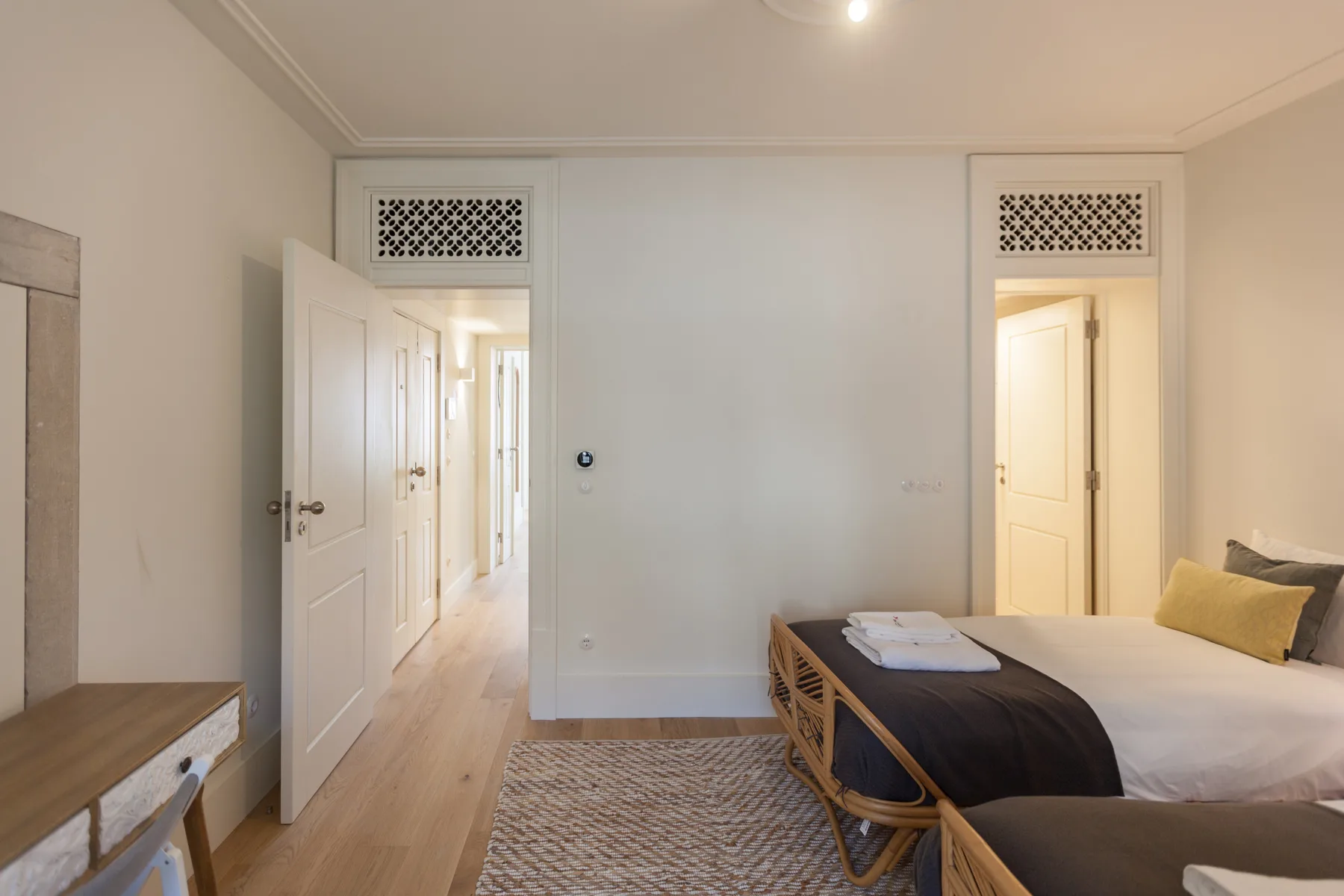 Dormitorio con una cama, un banco de fibras trenzadas y puertas abiertas a un pasillo.